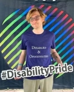 Nico is wearing a blue/purple shirt that says Disabled and Opinionated and holds a sign that says #DisabilityPride in front of a colorful background.  He has blond and brown hair and wears glasses.