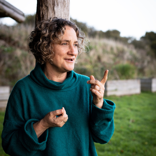 Woman with curly hair, wearing a teal turtleneck sweater, outdoors near a tree, gesturing with her right hand.