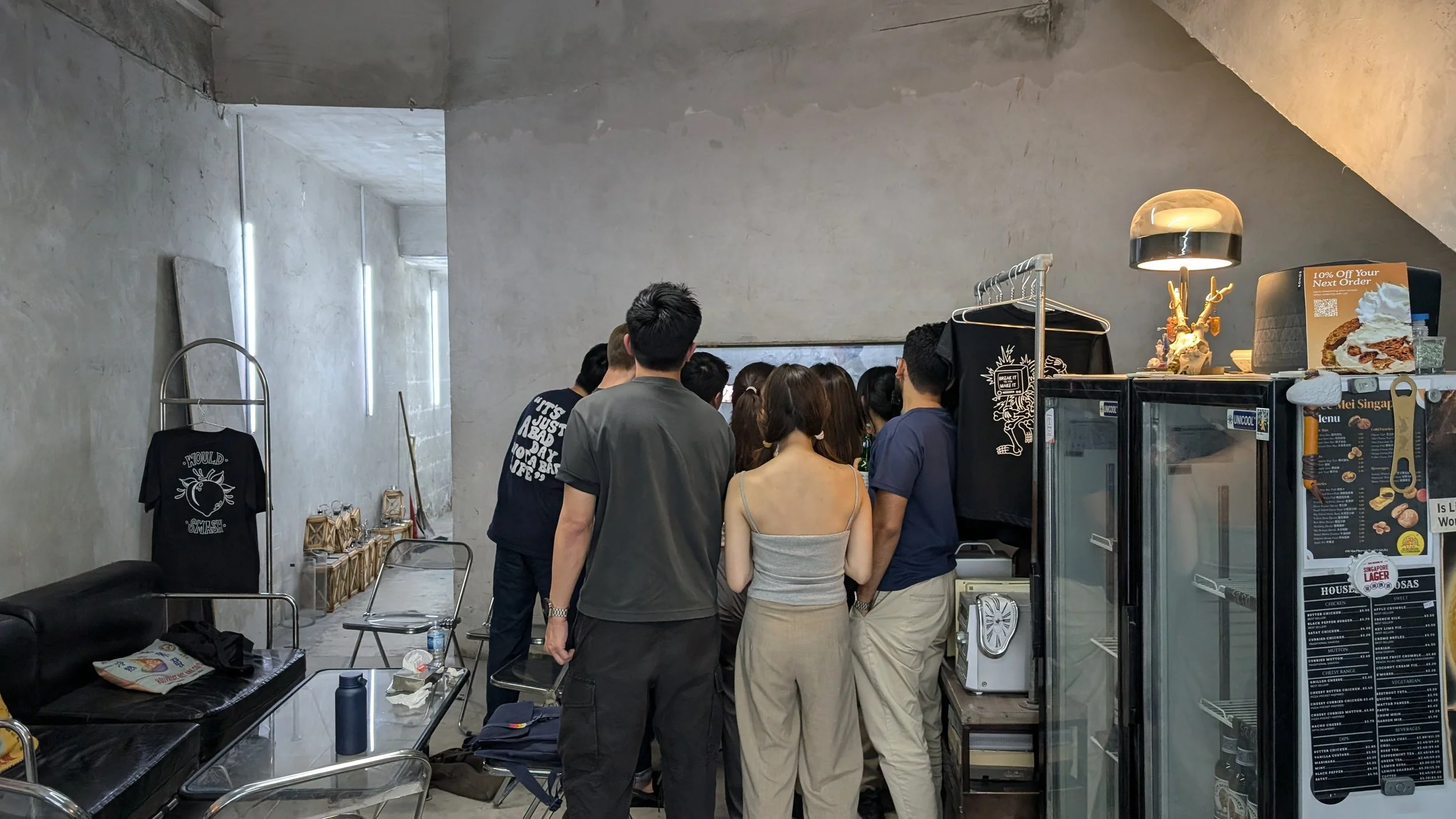 Group of people gathered closely together inside a cafe, standing around the counter area. The space has concrete walls and minimal decor, with some merchandise on display to the right and seating on the left.