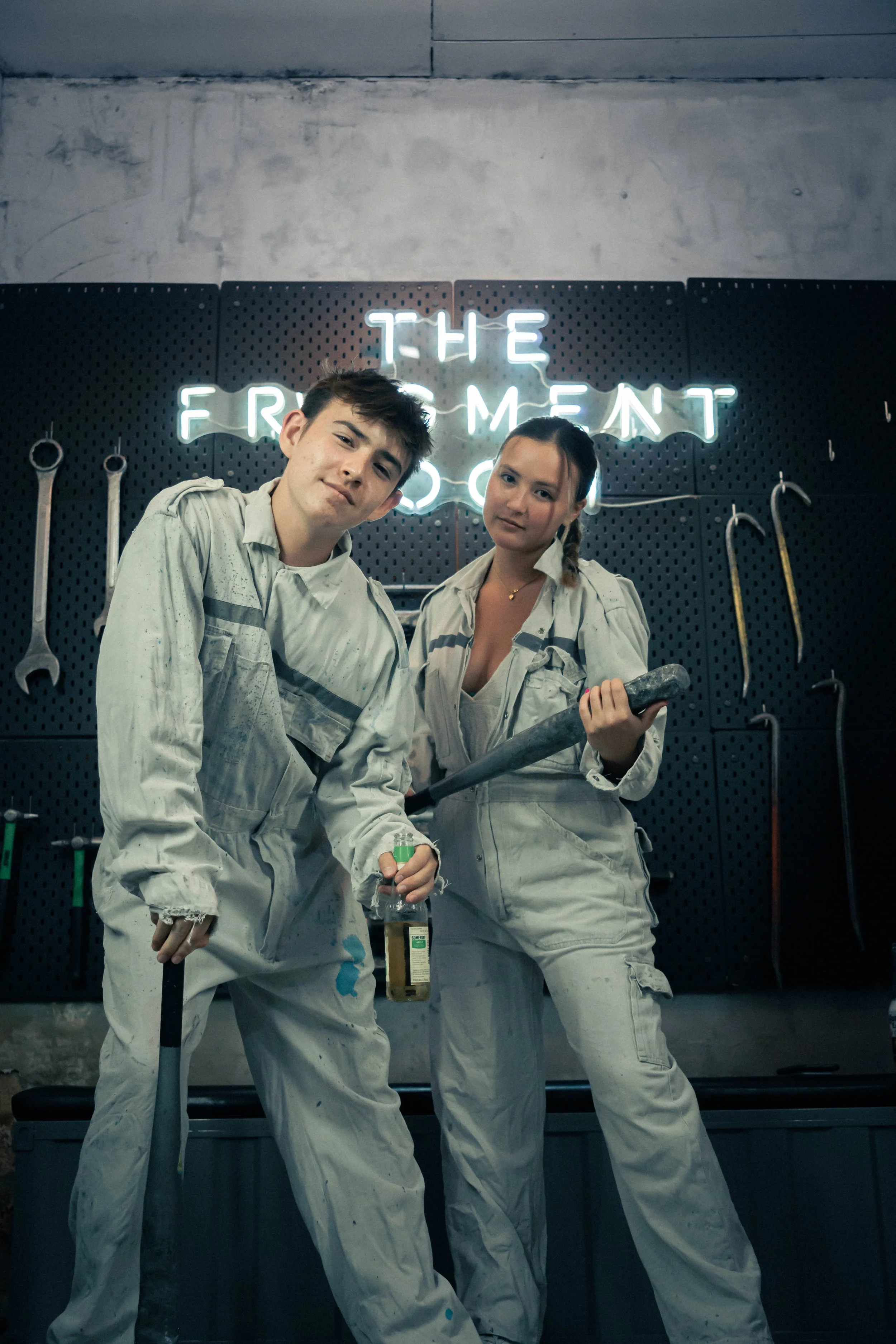 Two people in work overalls standing in front of a black pegboard wall with various tools hanging, with a neon sign behind them reading 'The Frightenment'.