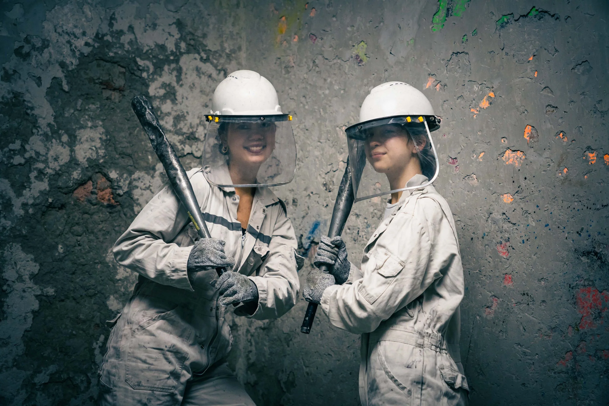 Two women in mining helmets and overalls holding sledgehammers, standing against a textured rock wall.