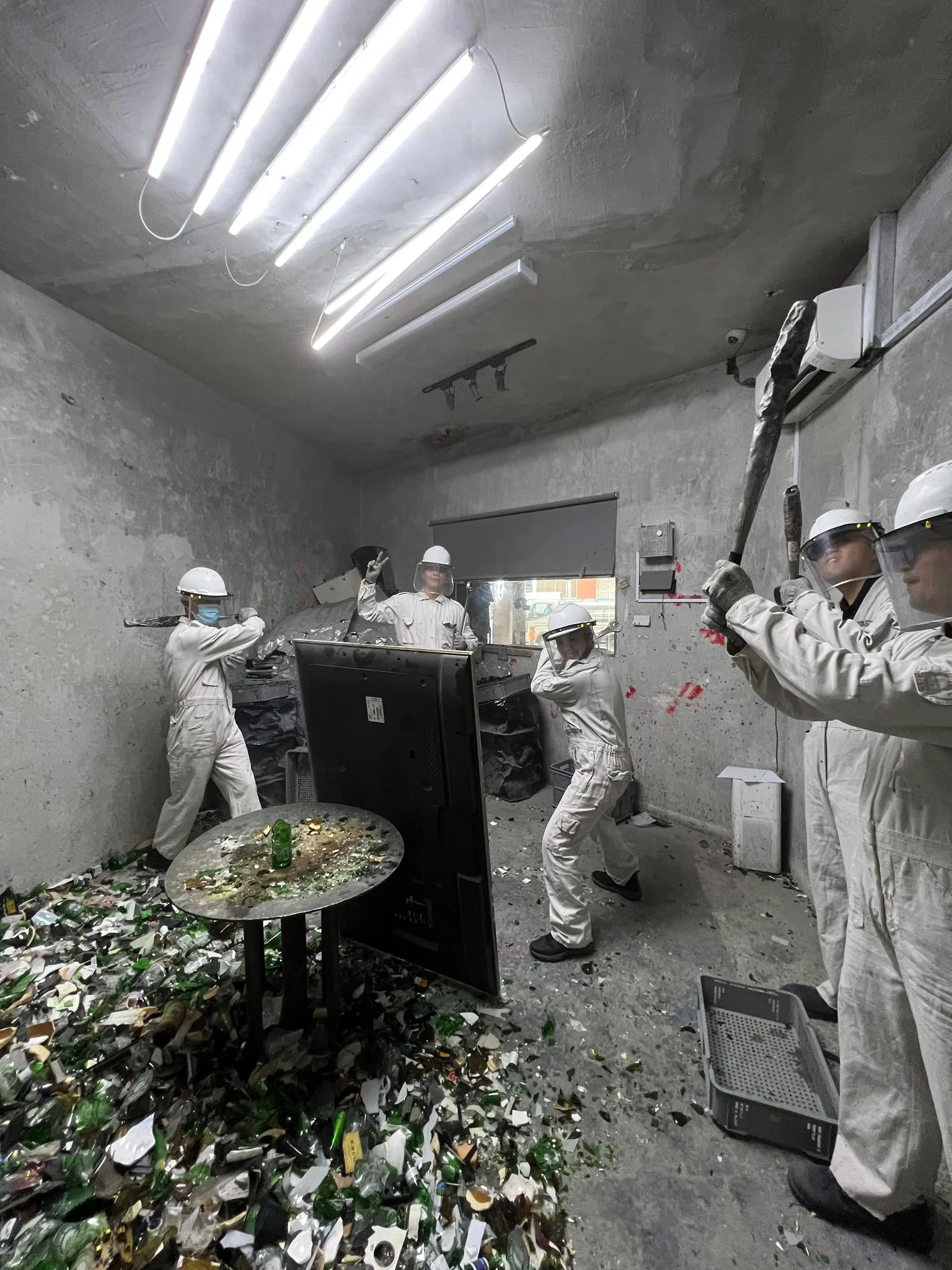 A group of workers dressed in white protective suits, helmets, and face shields are smashing electronic waste, such as broken glass and bottles, with tools inside an industrial room with gray walls and fluorescent lighting.