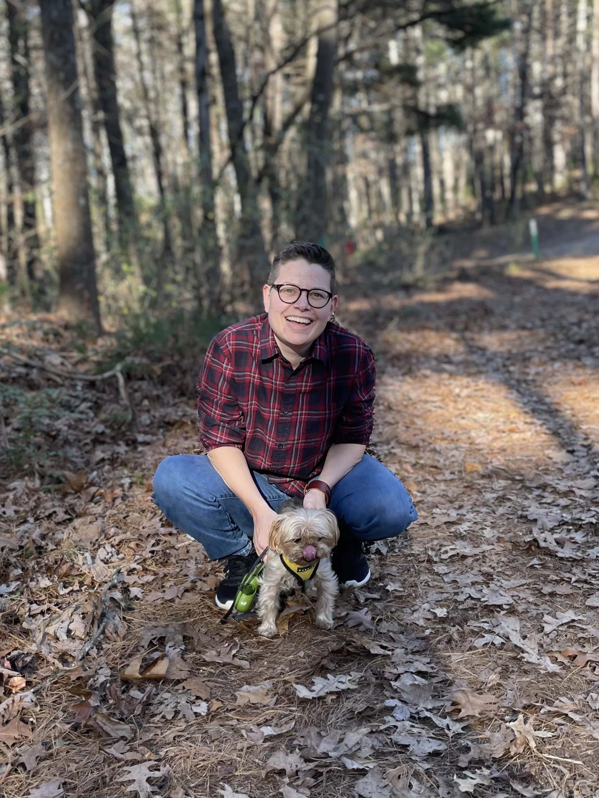 A person wearing glasses, a red plaid shirt, and blue jeans is squatting on a wooded trail, smiling at the camera. A small dog wearing a yellow harness and leash is in front of them, panting with its tongue out. The background features leafless trees and a dirt path covered with fallen leaves.