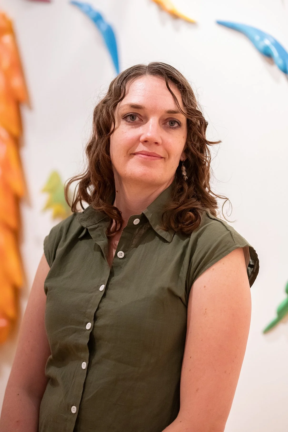 A woman with light skin, curly brown hair, wearing a green short-sleeve button-up shirt, standing indoors with colorful decorations in the background.