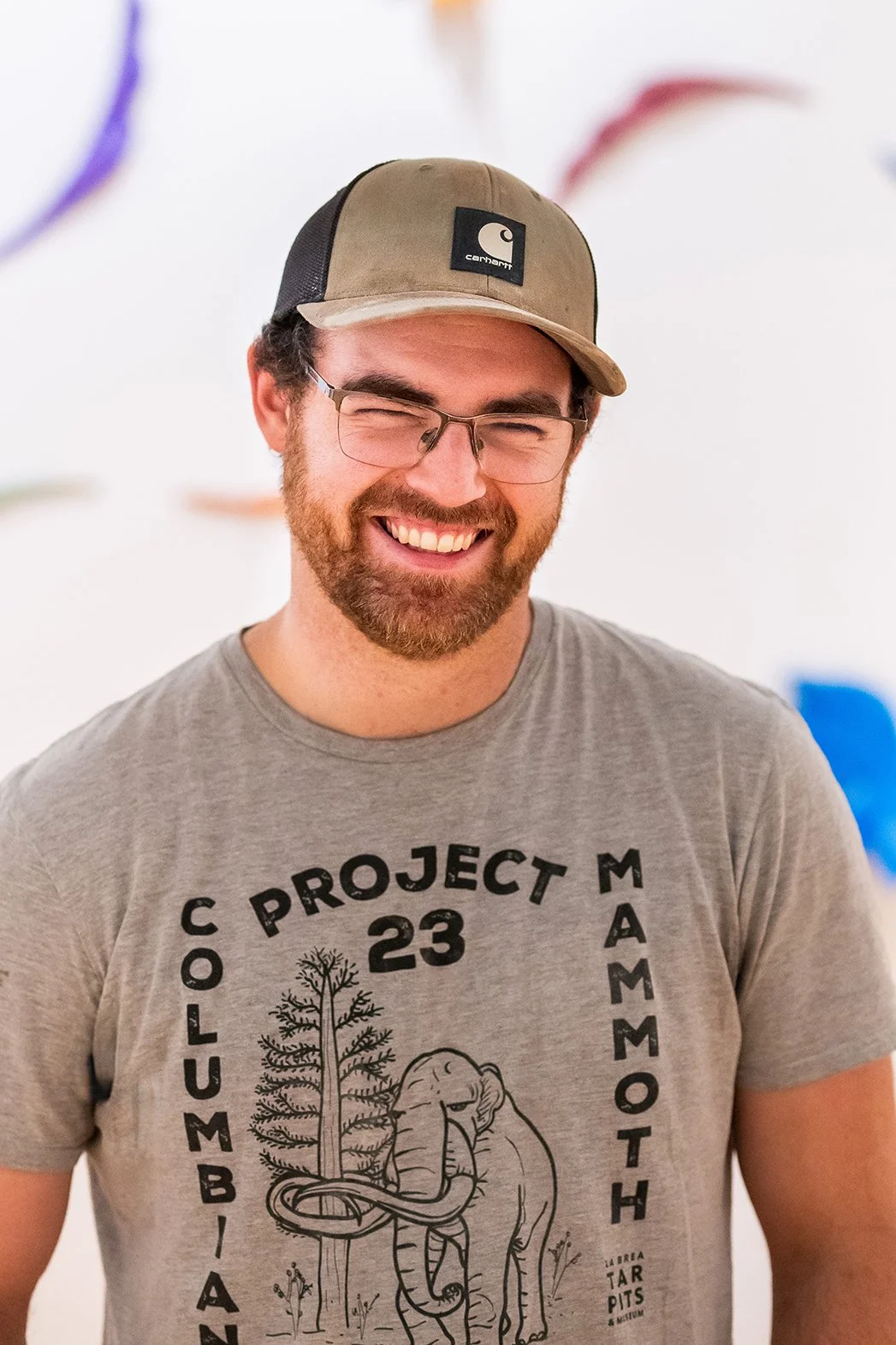 A smiling man with glasses, a beard, and a mustache wearing a tan and black Carhartt cap and a gray T-shirt with text and a drawing of an elephant, trees, and a snake.