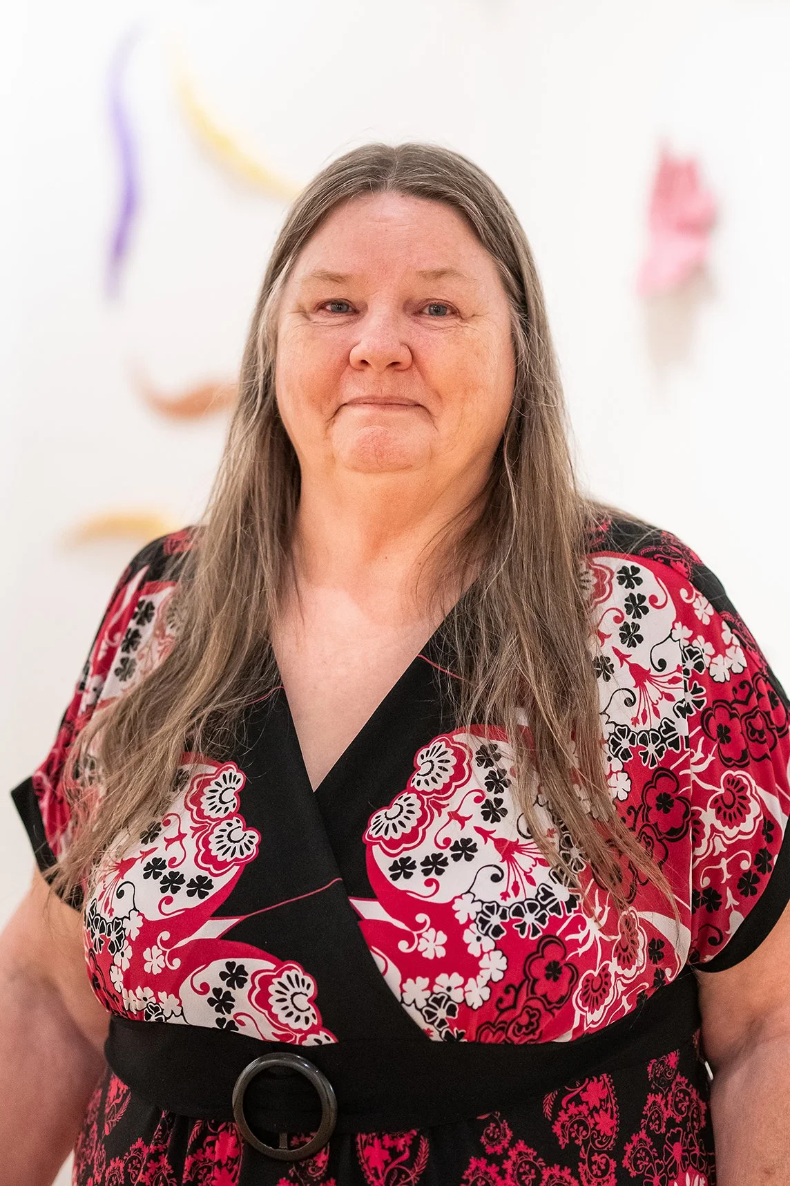 A woman with long hair wearing a black and red floral dress standing in front of an art installation with colorful fabric sculptures hanging on a white wall.