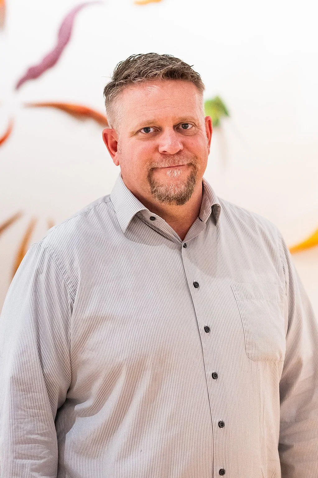A middle-aged man with short, styled hair and a goatee, wearing a light-colored, button-up dress shirt, standing indoors against a white wall with colorful decor.