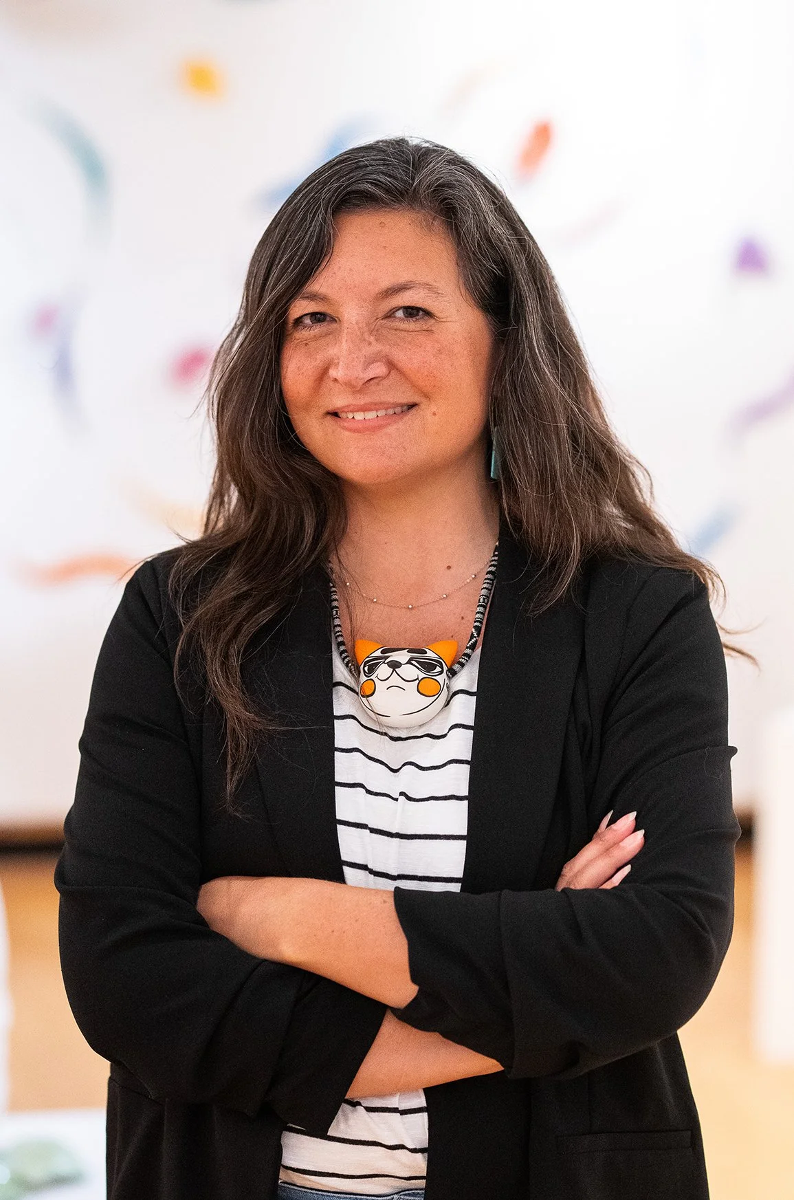 A woman with long dark hair wearing a black blazer over a striped shirt, standing with arms crossed, smiling, with colorful blurred shapes in the background.