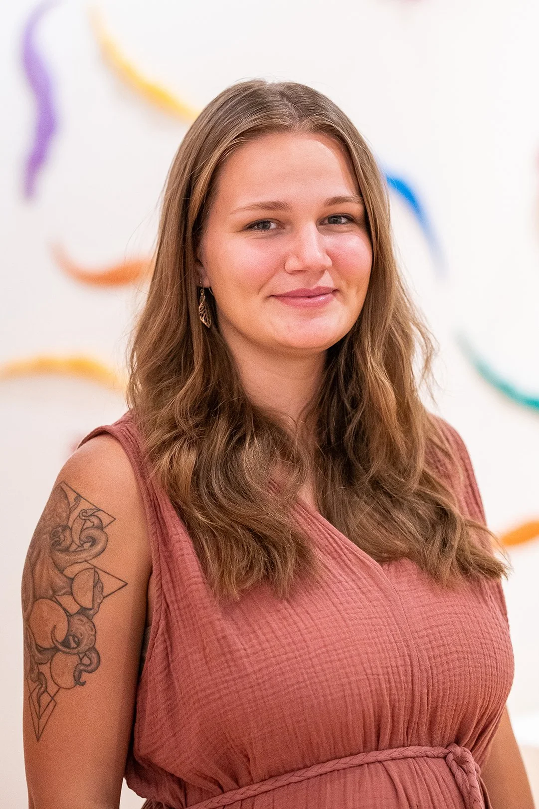 A smiling young woman with long, wavy brown hair and a tattoo on her upper arm, wearing a sleeveless pinkish top, standing in front of a white wall with colorful abstract art.