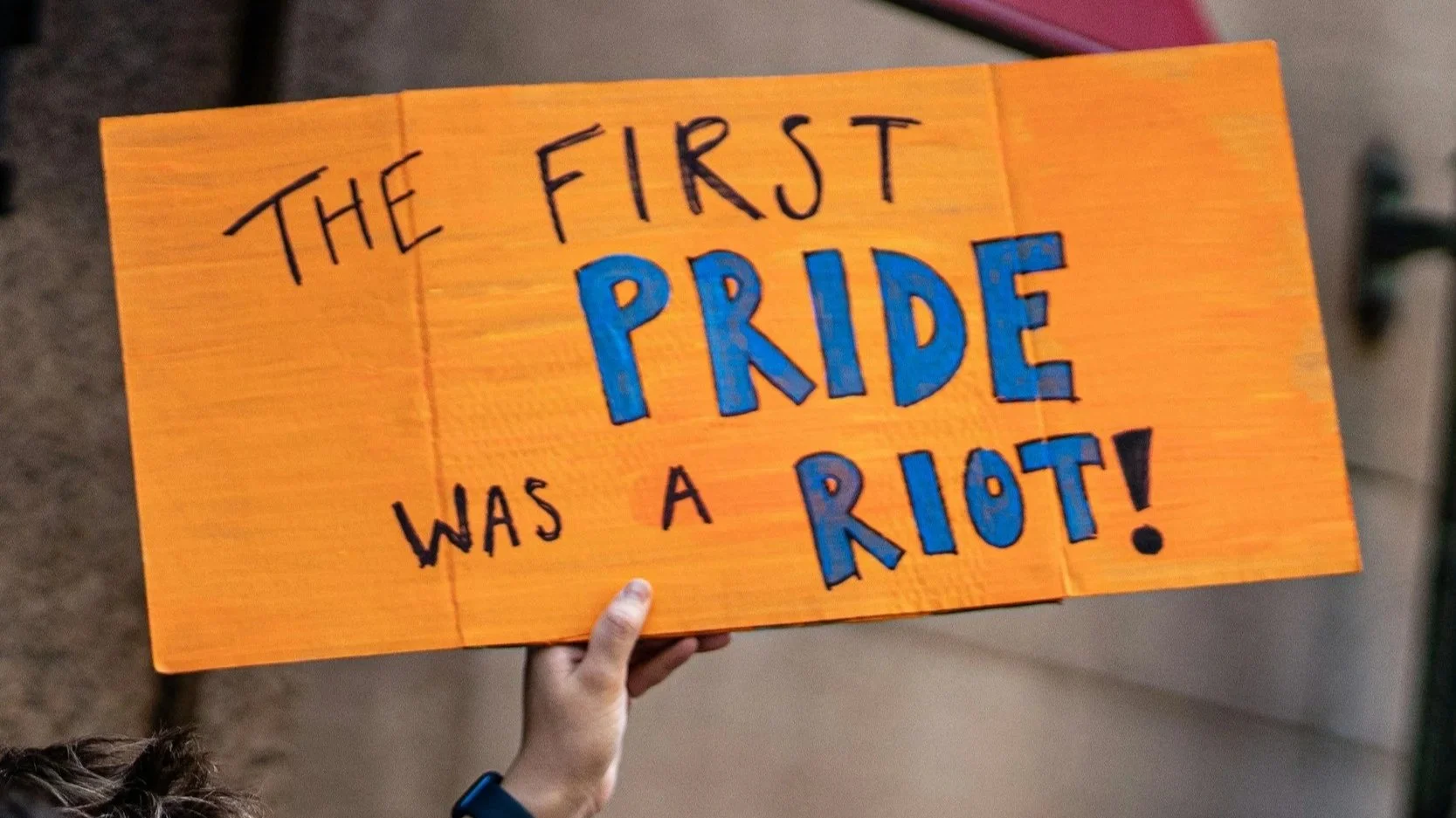 Person holding a sign reading "The First Pride Was a Riot," highlighting the Stonewall Uprising and the history of Pride Month.