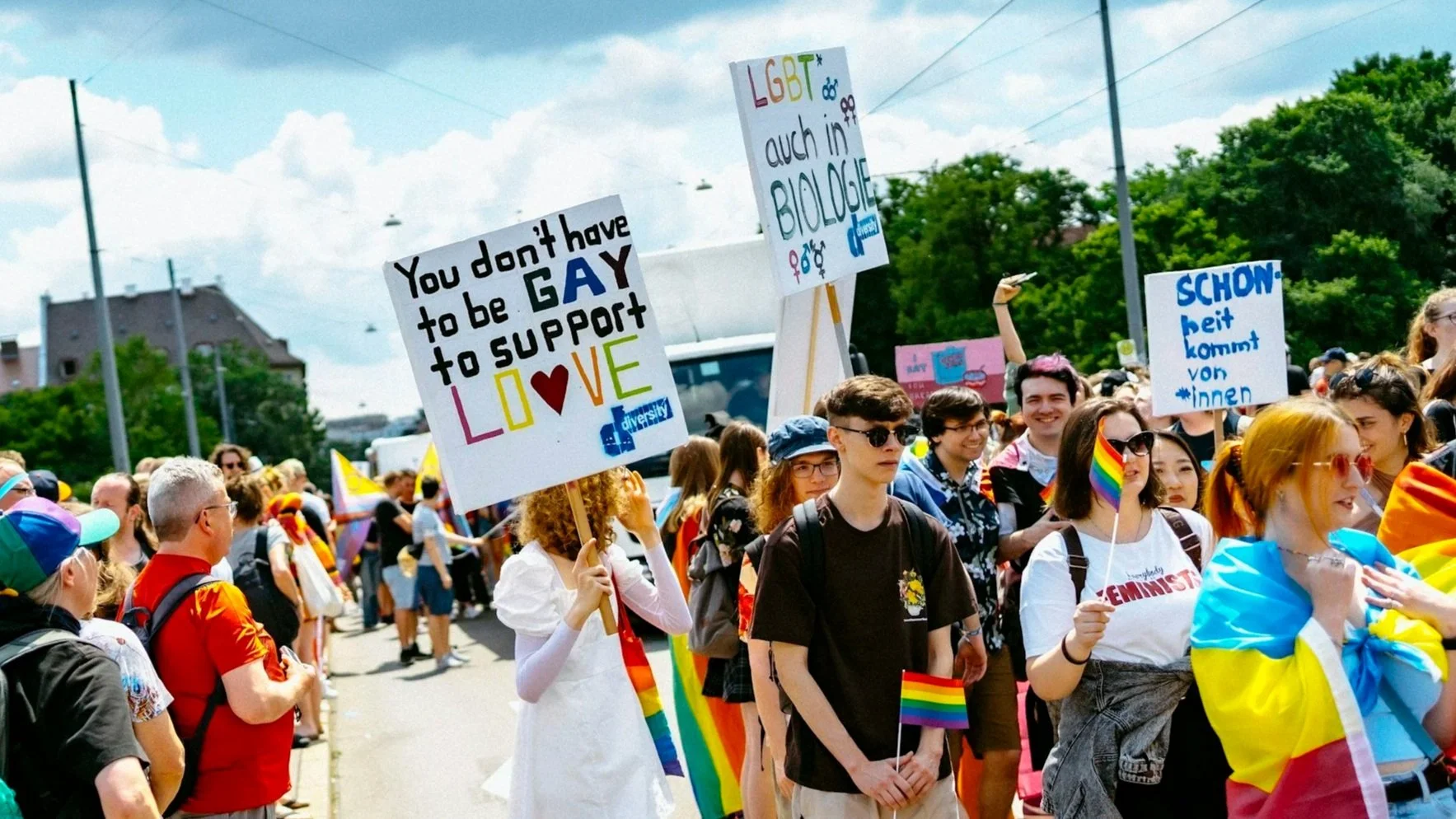 Pride Month attendees marching with a sign reading "You don't have to be gay to support LOVE," highlighting allyship and support for the LGBTQIA+ community.