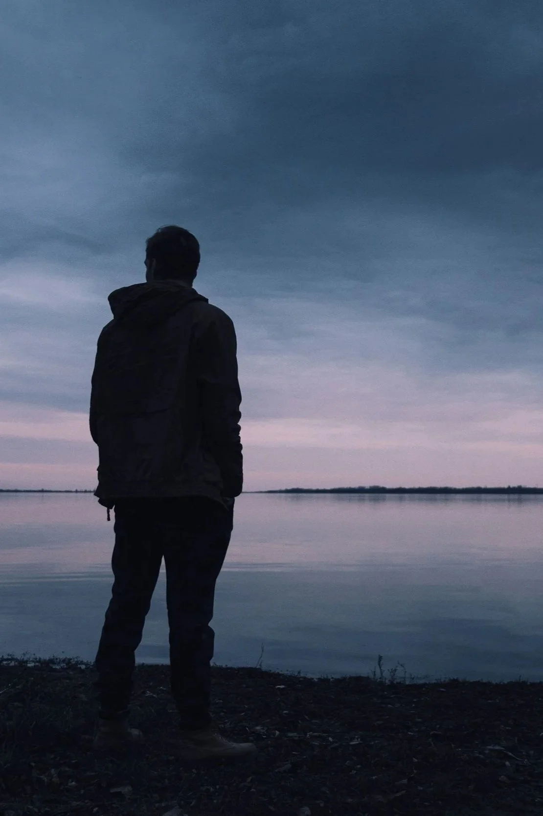 Silhouetted man standing on a lakeshore at dusk.