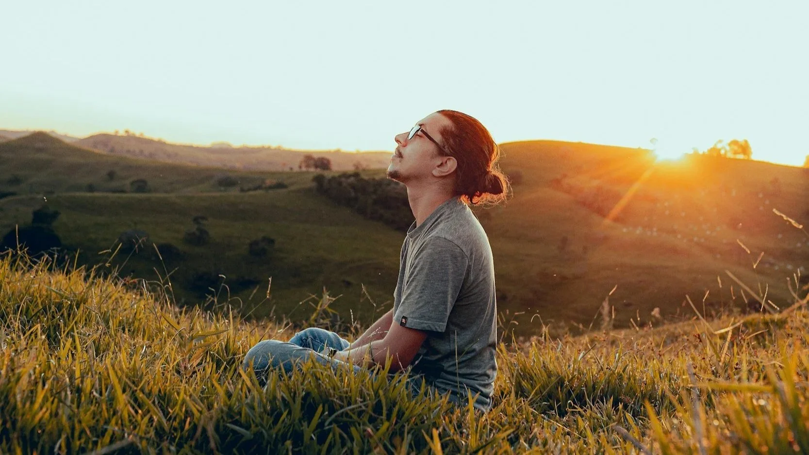 A man sitting cross-legged in a green country side with rolling hills, the sun is rising in the background.
