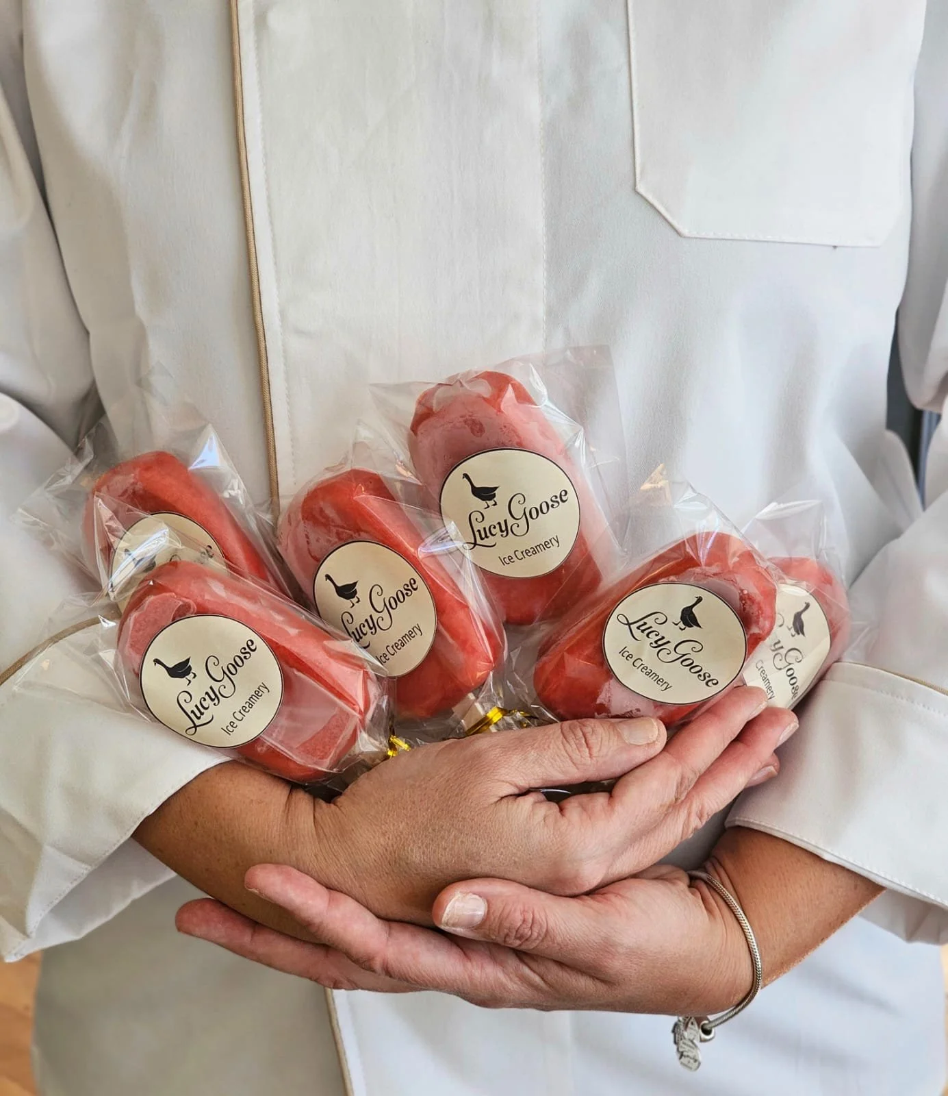 A person in a white apron holding multiple pink ice cream bars wrapped in clear plastic with a Lucy Goose Ice Creamy label.