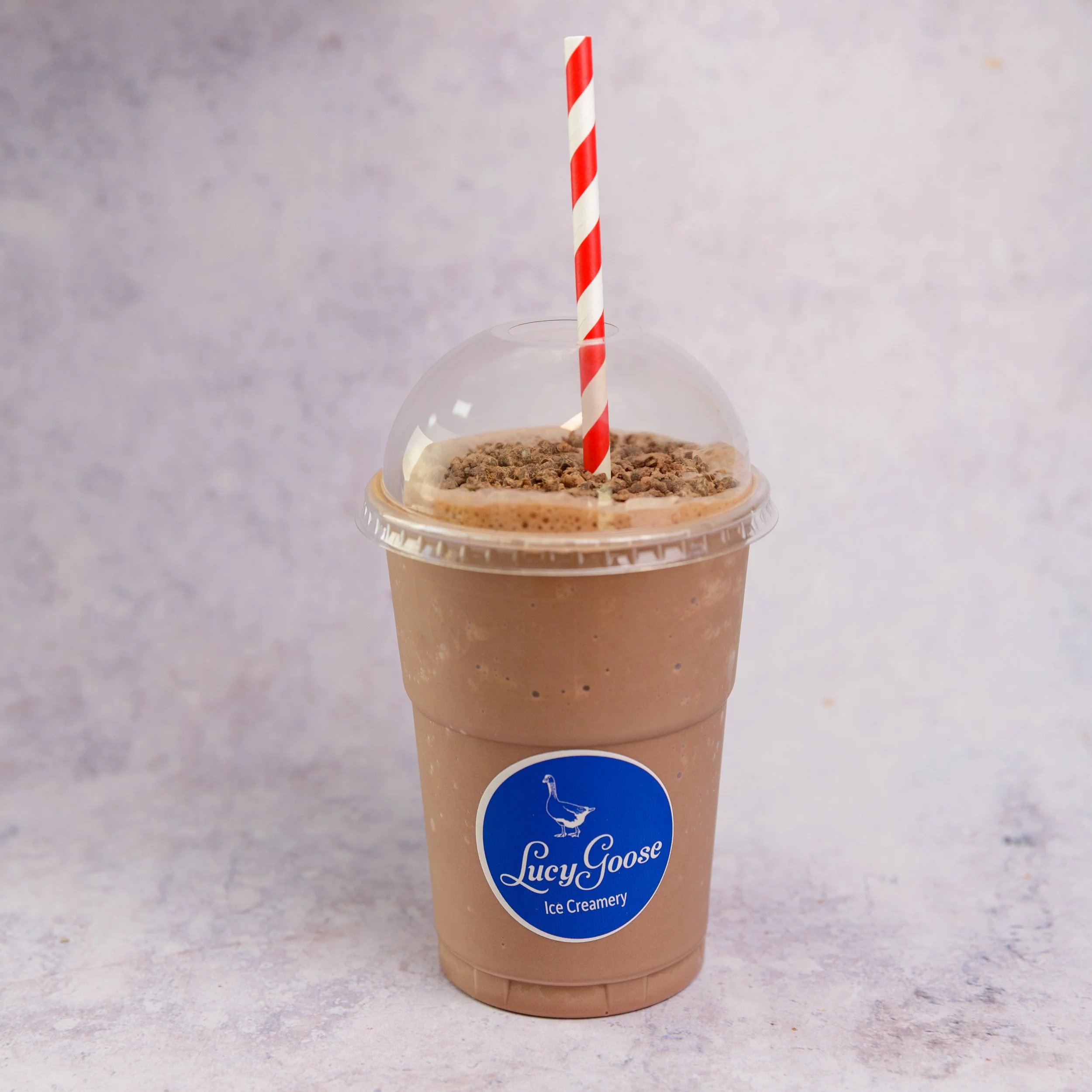 A blended chocolate milkshake in a clear cup with whipped topping and chocolate sprinkles, topped with a striped paper straw, labeled Lucy Goose Ice Creamery.