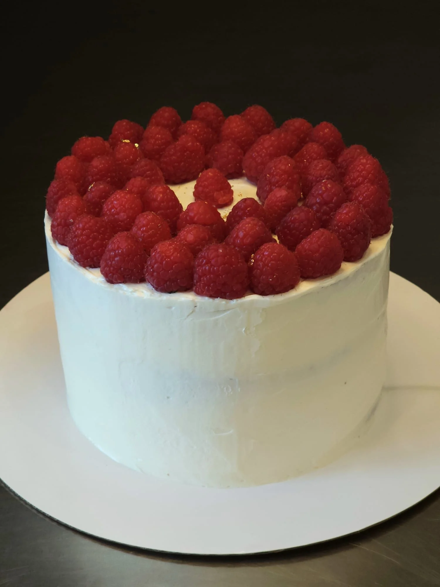 A round cake topped with fresh raspberries and cream, sitting on a white cake board against a dark background.