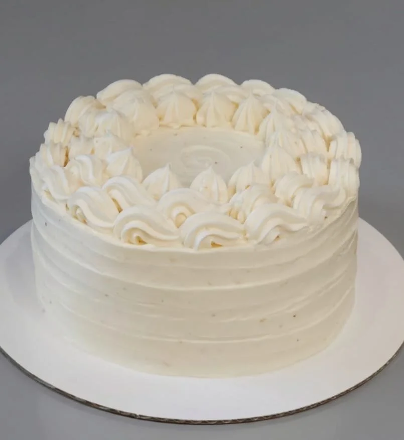 A round white cake with piped frosting decorations on top, placed on a white cake board against a gray background.