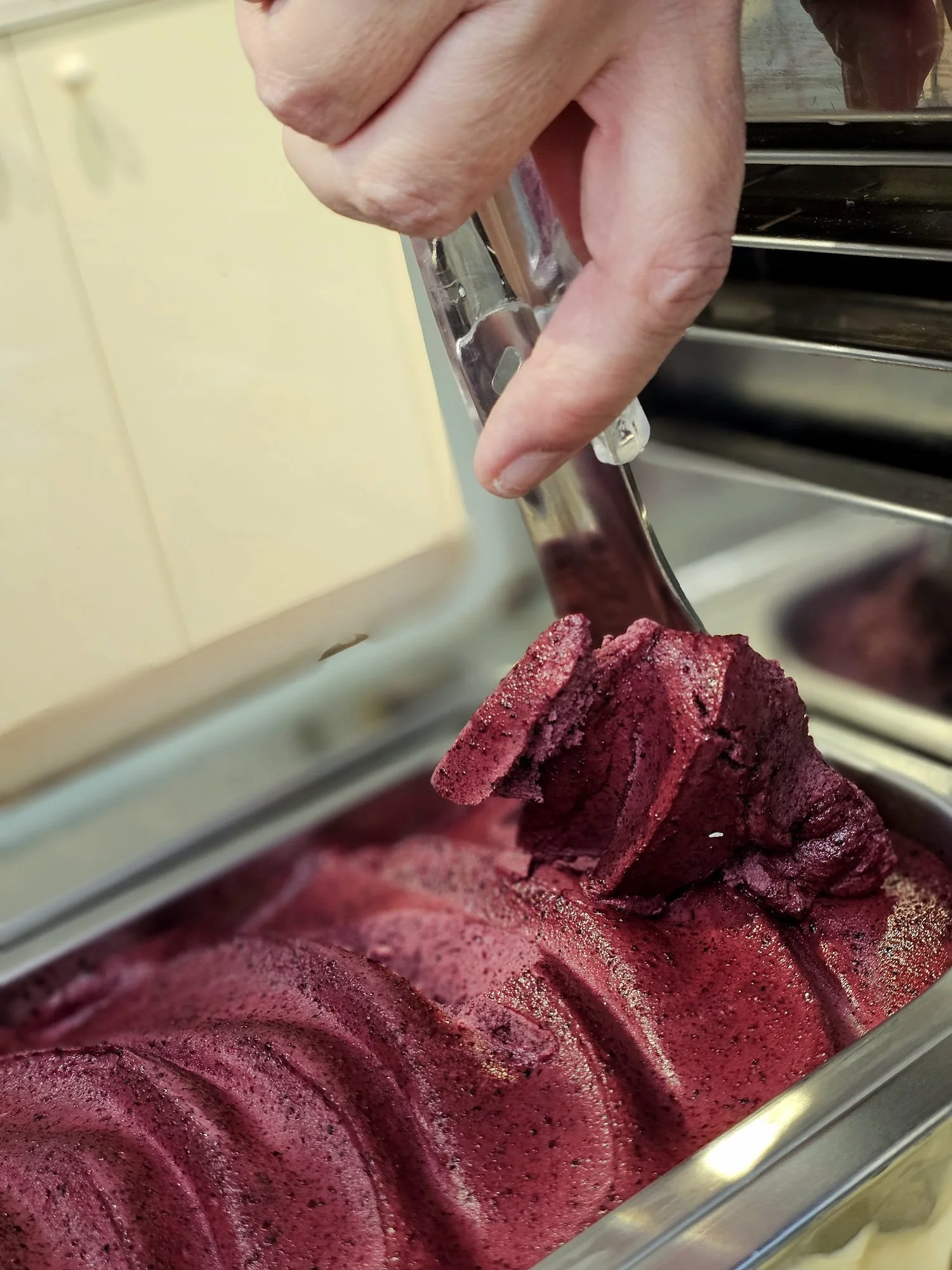 A person's hand using ice cream tongs to scoop reddish-purple sorbet or ice cream from a metal container.