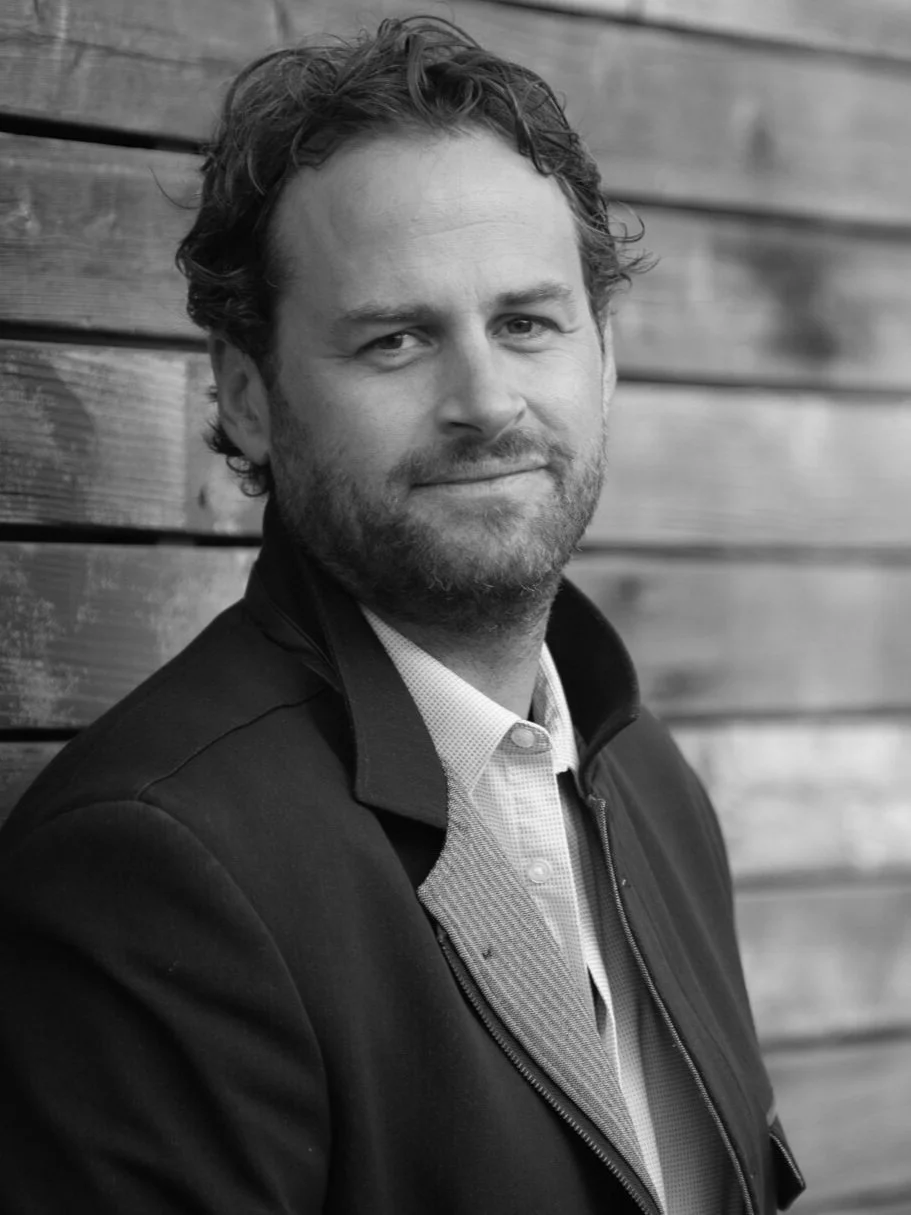 Retrato en blanco y negro de un hombre con barba, cabello rizado y expresión amable, vestido con chaqueta y camisa, posando junto a una pared de madera.