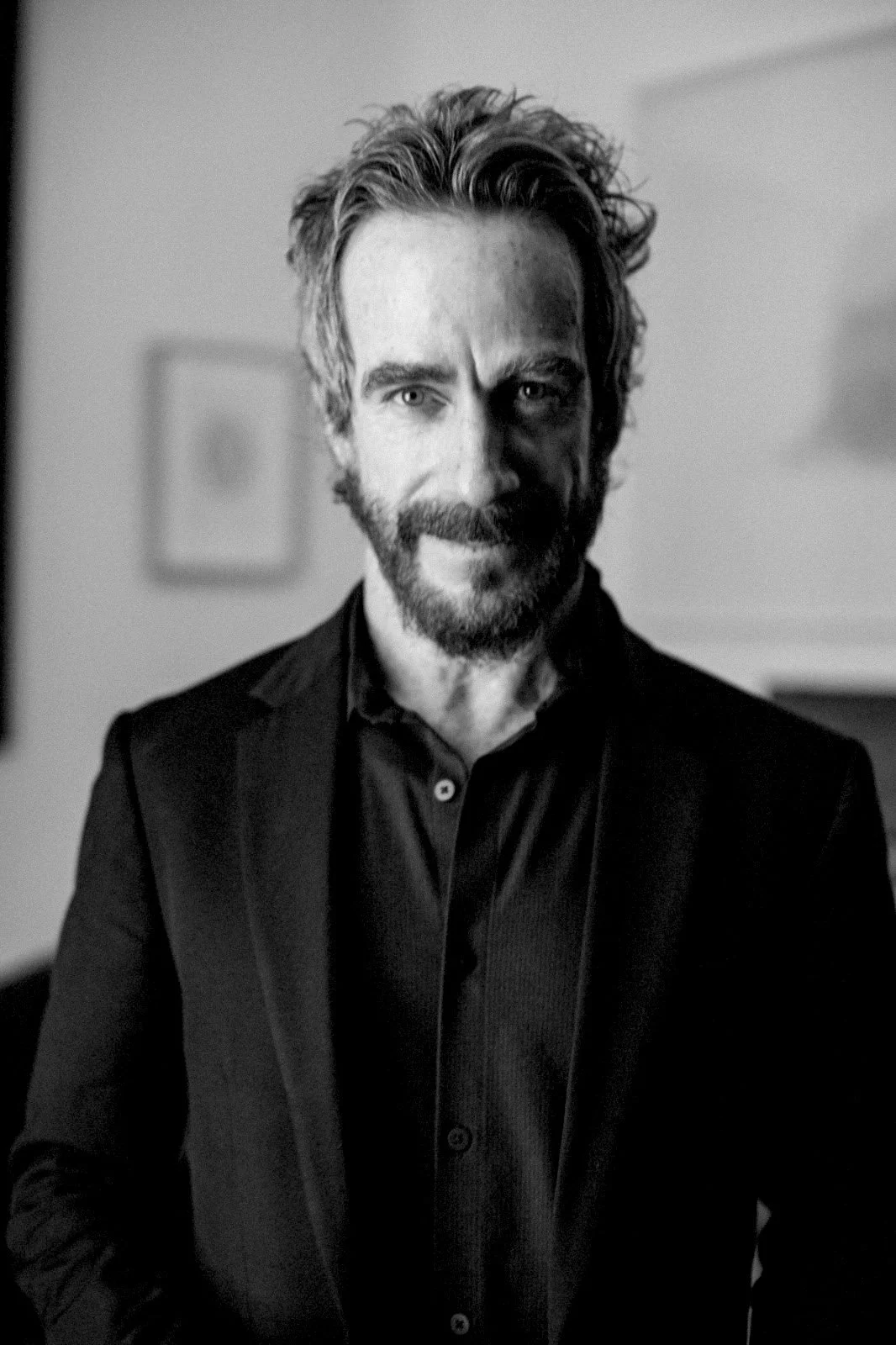 Retrato en blanco y negro de un hombre con barba, cabello rizado y expresión confiada, vestido con chaqueta y camisa oscuros, en un entorno interior.