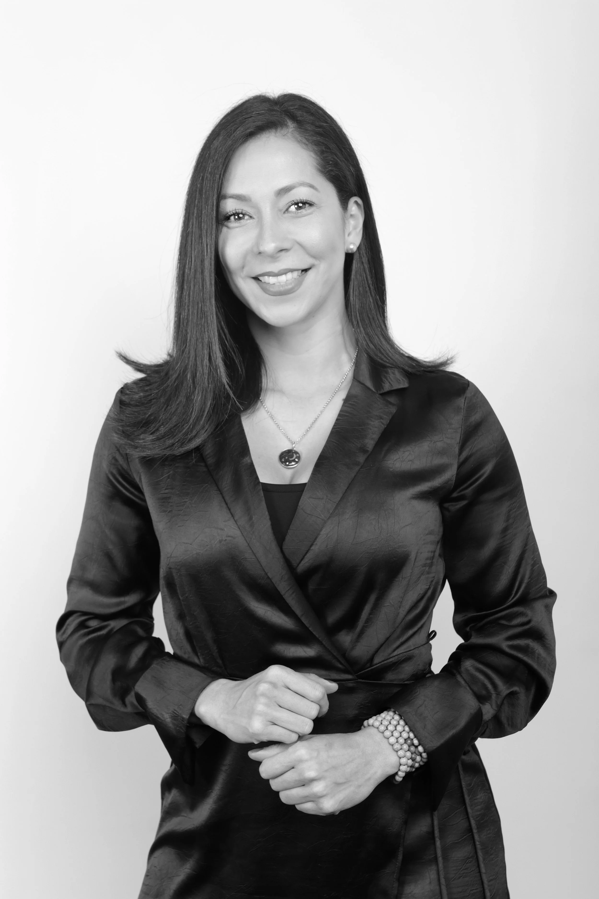 Retrato en blanco y negro de una mujer con cabello largo y liso, sonriendo, usando un blazer de satén, collar y pulsera de perlas, con fondo blanco.