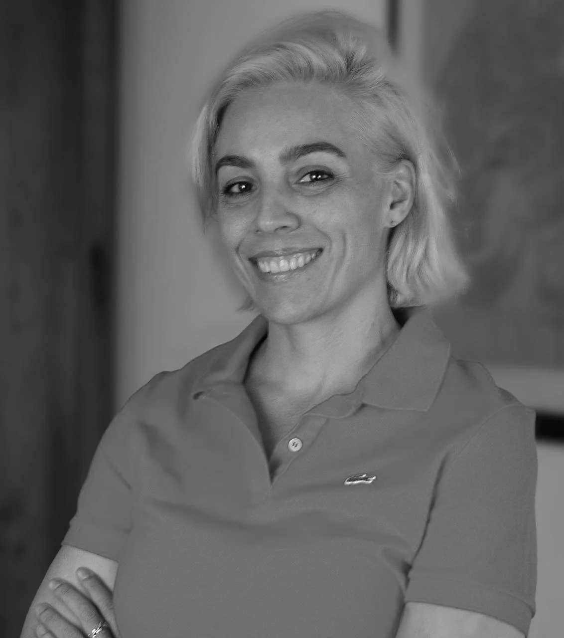 Retrato en blanco y negro de una mujer con cabello rubio corto, sonriendo y con los brazos cruzados, usando una camiseta polo.
