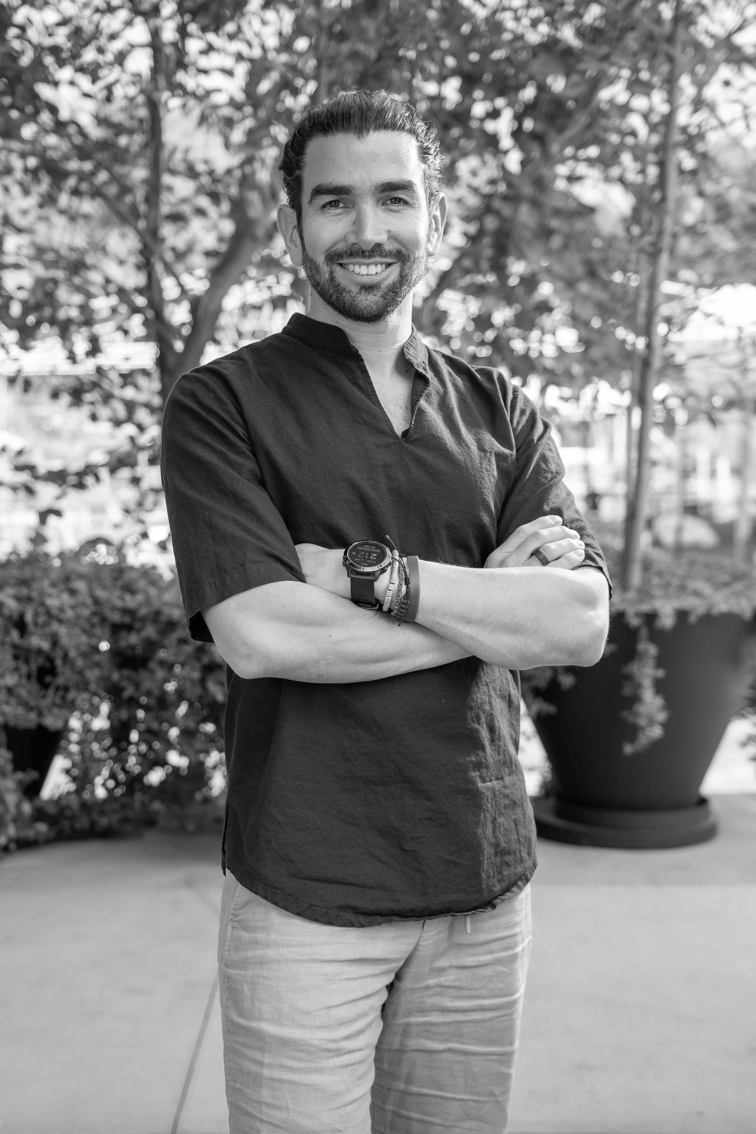 Hombre sonriendo, con barba y cabello recogido, de brazos cruzados, en un entorno con plantas y árboles, en blanco y negro.