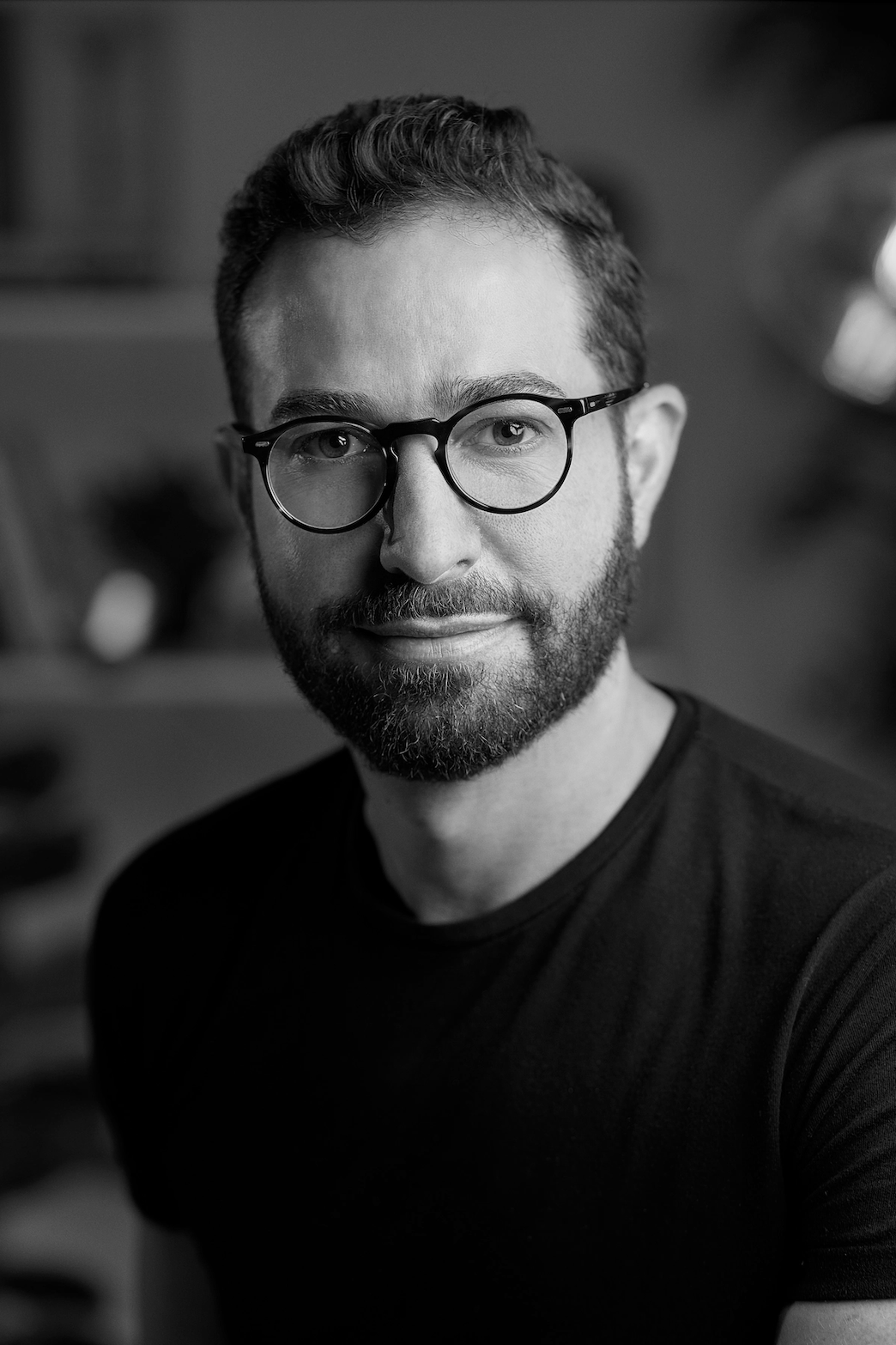 Retrato en blanco y negro de un hombre con barba, usa gafas de montura redonda y camiseta negra, sonriendo ligeramente.
