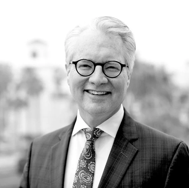 Hombre de cabello canoso, con gafas negras, vestimenta formal con saco y corbata, sonriendo en un fondo borroso en escala de grises.