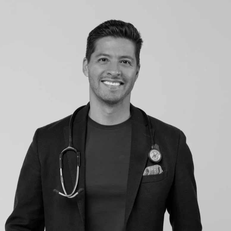 Hombre con cabello oscuro, sonriendo, lleva una bata de médico y un estetoscopio colgado del cuello.