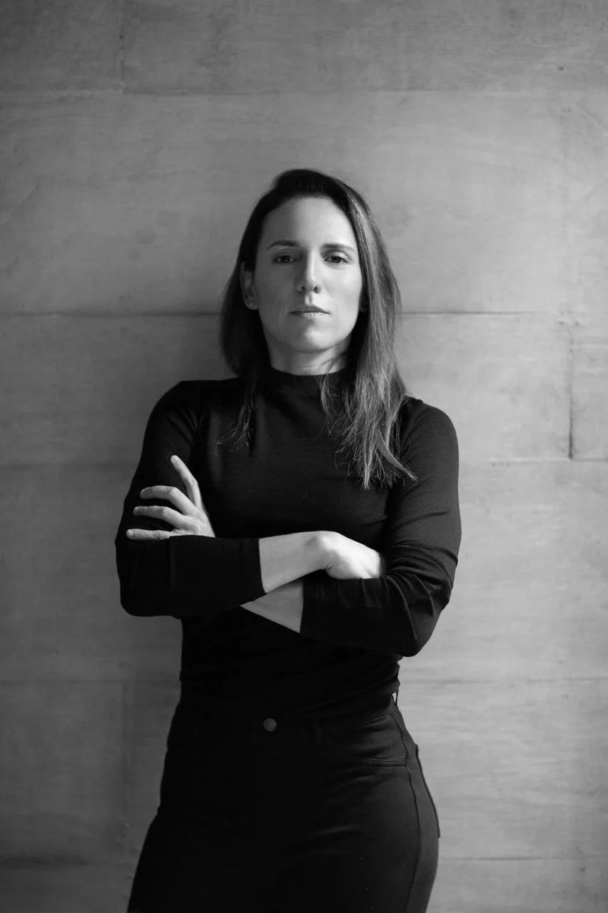 Retrato en blanco y negro de una mujer con cabello largo y oscuro, vestida con camisa de manga larga y pantalones oscuros, de pie con los brazos cruzados frente a una pared de madera.