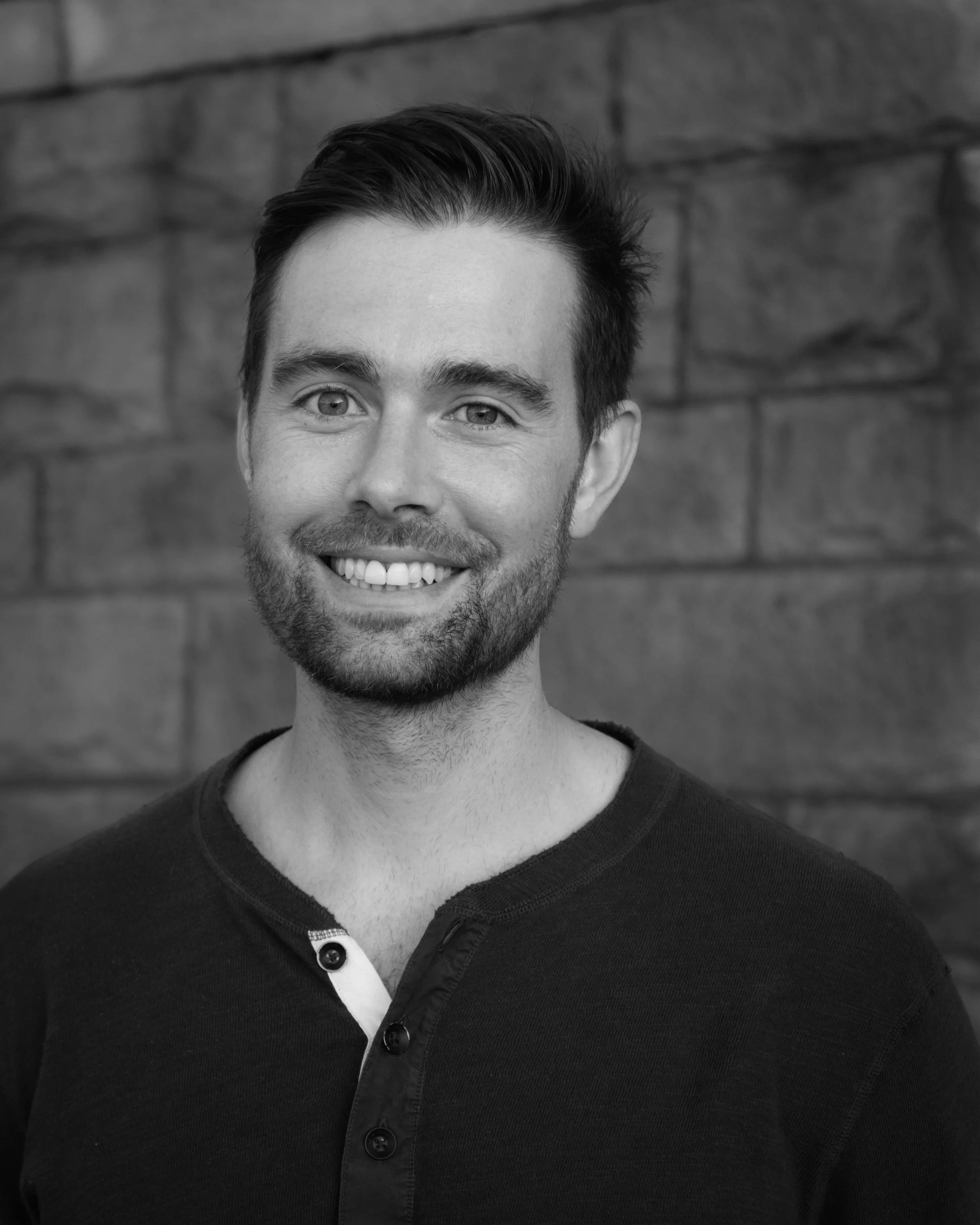 Retrato en blanco y negro de un hombre joven, sonriendo, con barba y cabello oscuro, con fondo de muro de ladrillo.