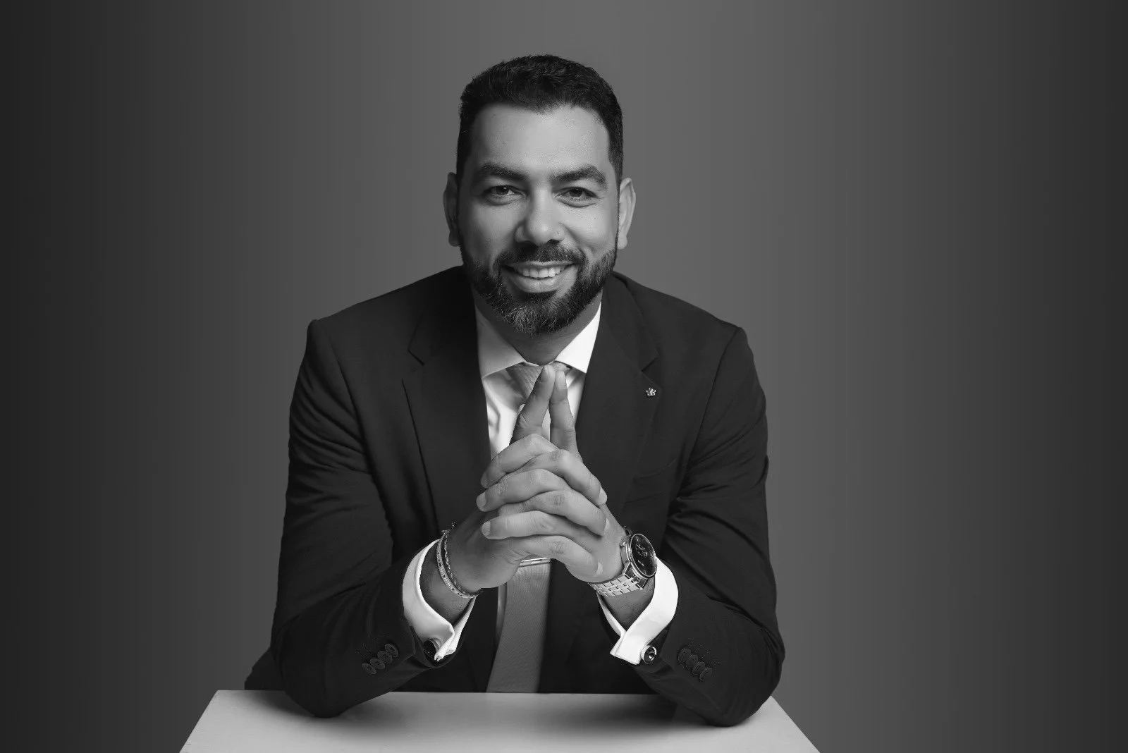 Retrato en blanco y negro de un hombre de aspecto profesional, con barba y cabello corto, vestido con traje y corbata, sonriendo con las manos juntas sobre una mesa.