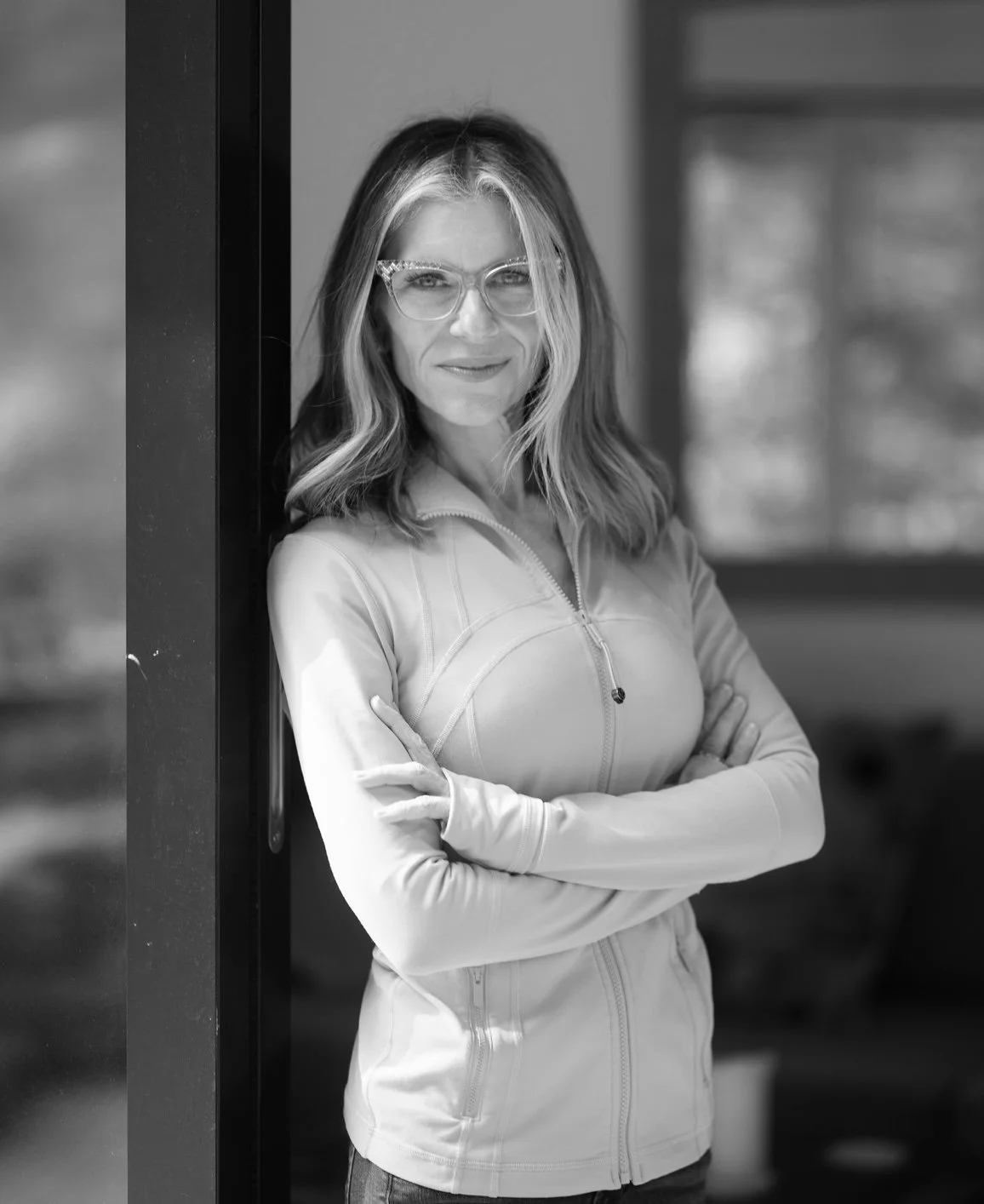 Mujer con gafas, cabello rubio y oscuro, usando chaqueta deportiva, posando con brazos cruzados y sonriendo ligeramente, en un entorno interior con ventanas.