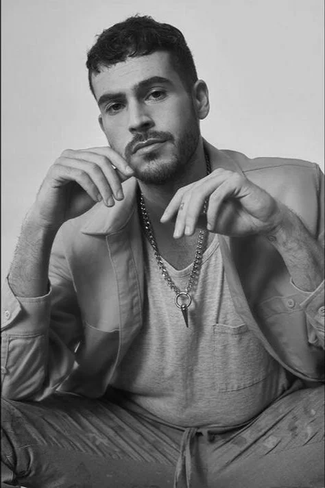Retrato en blanco y negro de un hombre joven con barba, usando una chaqueta y una camiseta, con cadenas, sentado con las piernas cruzadas y las manos cerca del rostro.