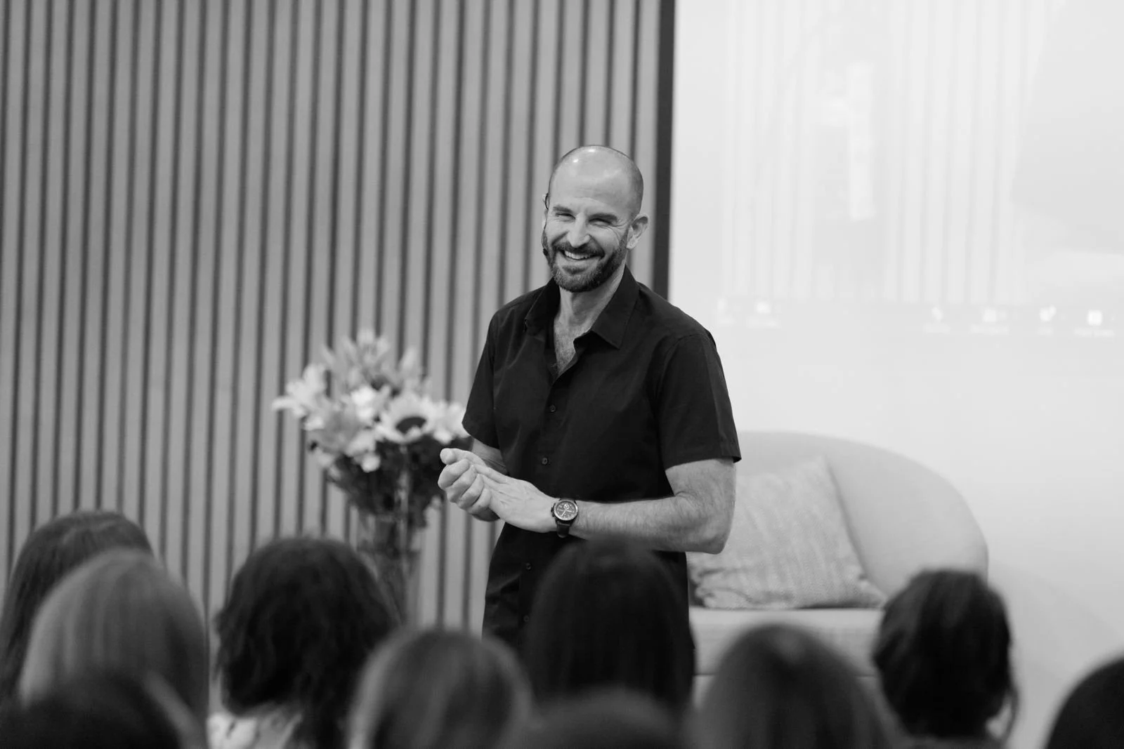 Hombre calvo con barba, sonriendo y vistiendo una camisa negra, dando una charla a un grupo de personas en un ambiente moderno.
