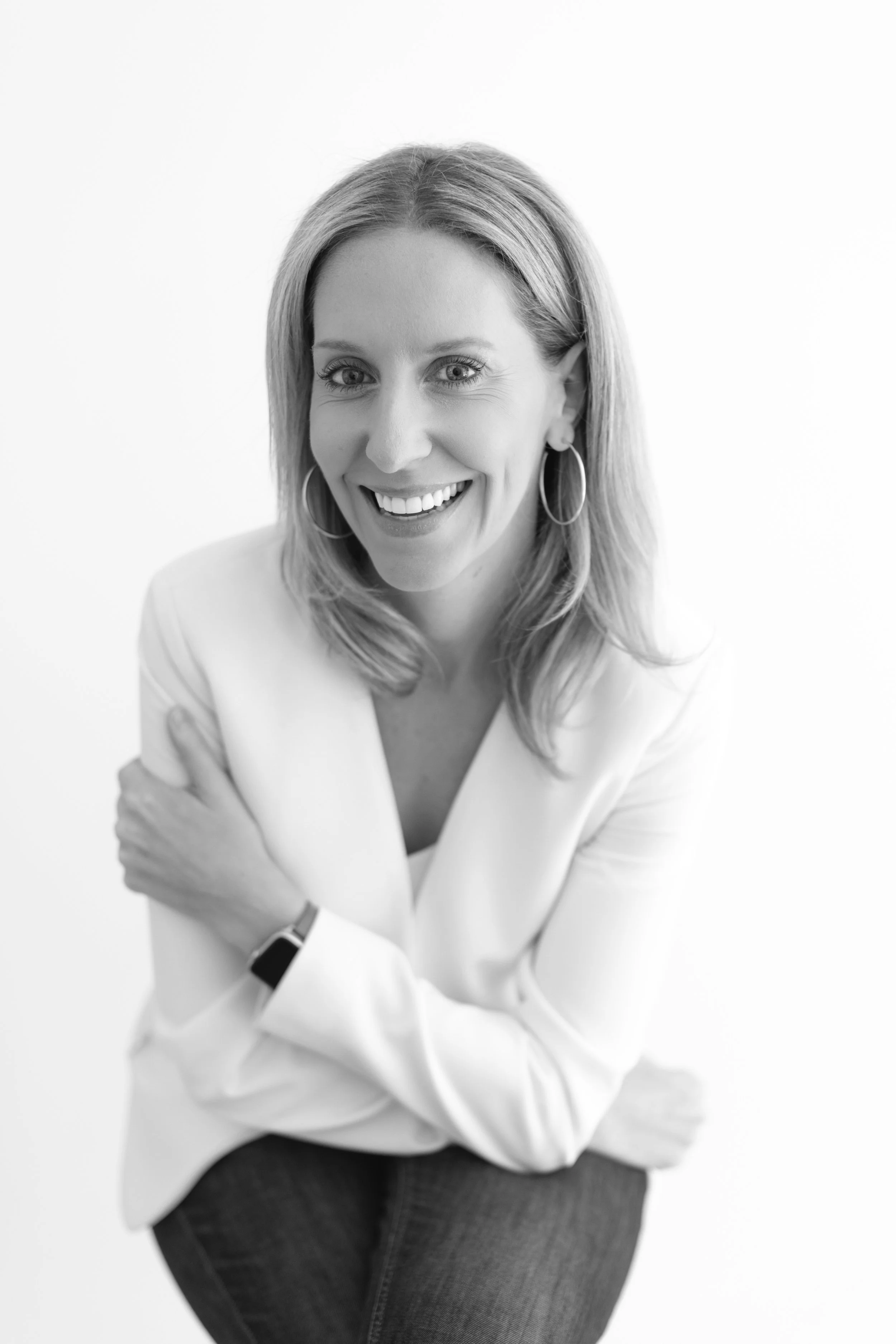 Mujer sonriendo, de cabello rubio y largo, usando una blusa blanca y aros, en un fondo blanco.