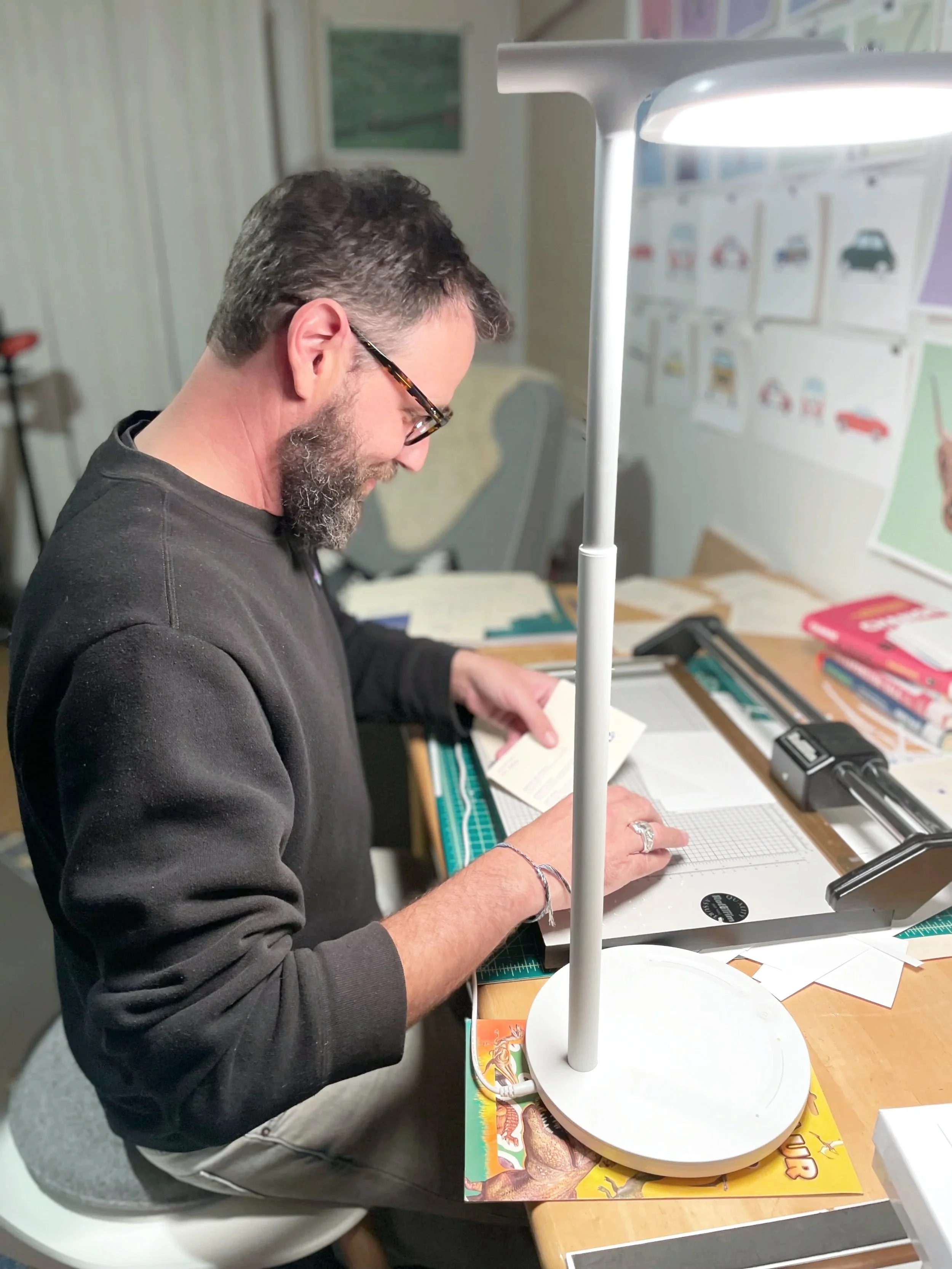 Image is a color photograph of the artist, Sam Slater, sitting at his desk in his studio. He is wearing glasses and has a salt and pepper beard.