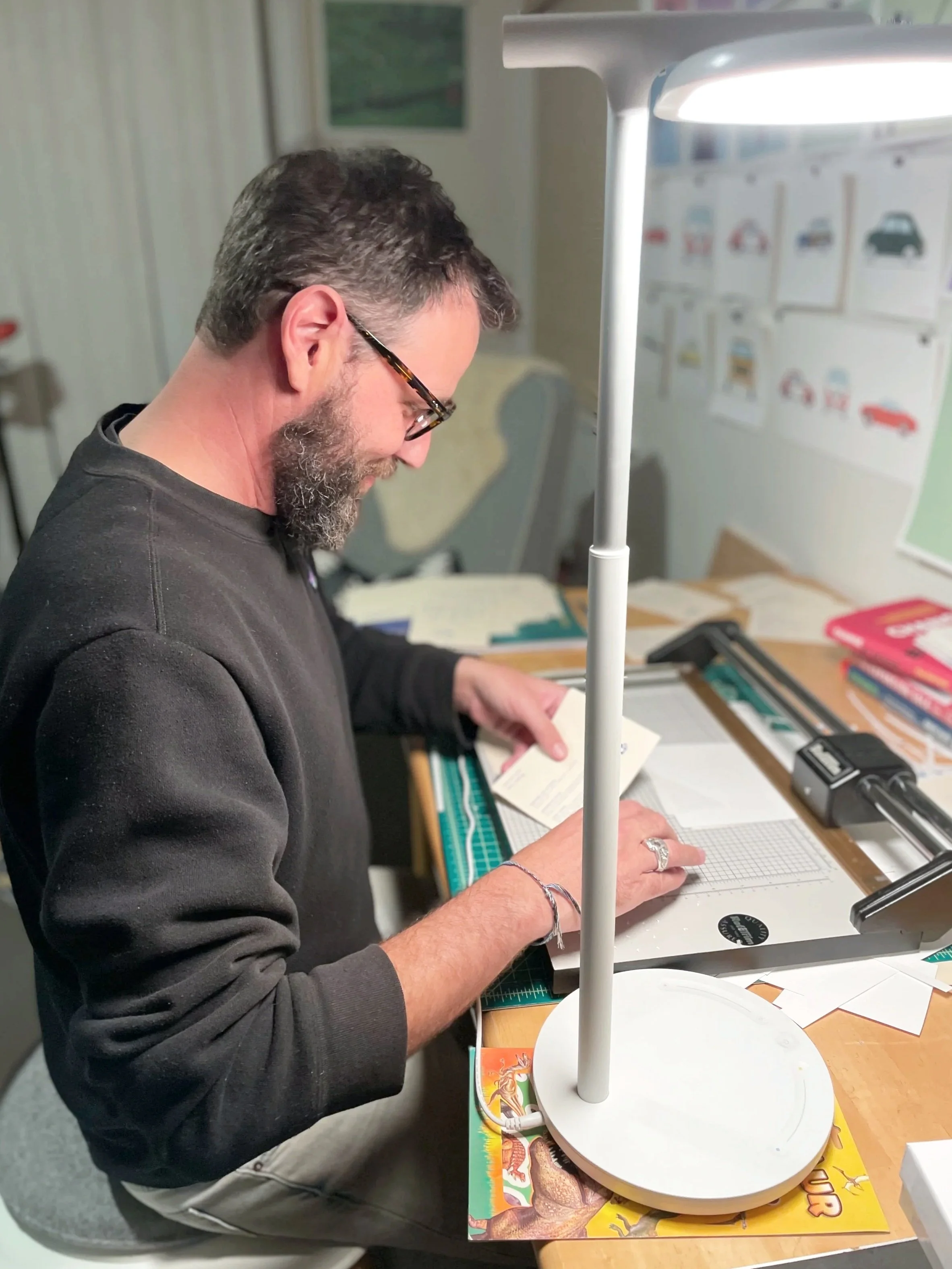 Sam Slater Art Studio. Picture is of a man with a beard and glasses, working at night in his studio. He is wearing a dark sweater. In the background you can see blurred images of his art work.