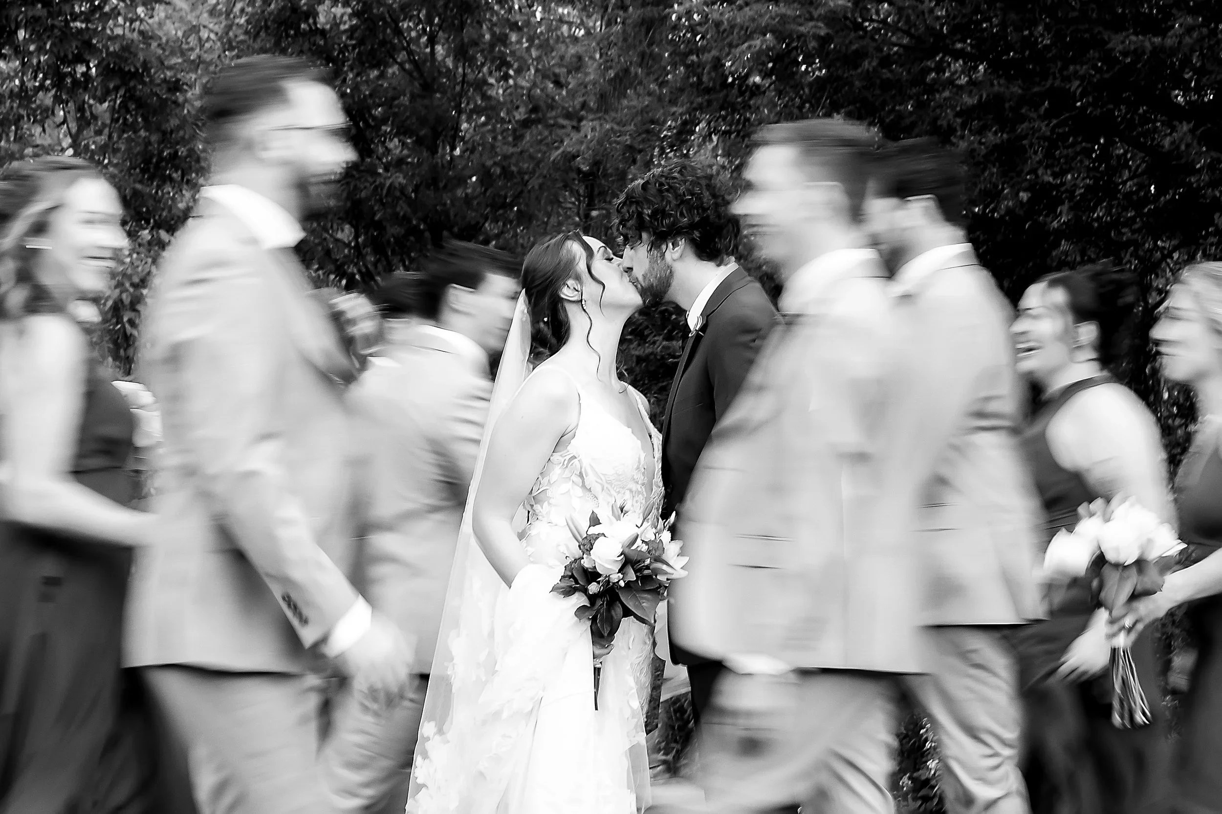 elegant black and white wedding photo of bride and groom kissing while the bridal party is blurred, moving around them, outside at Wellers Weddings