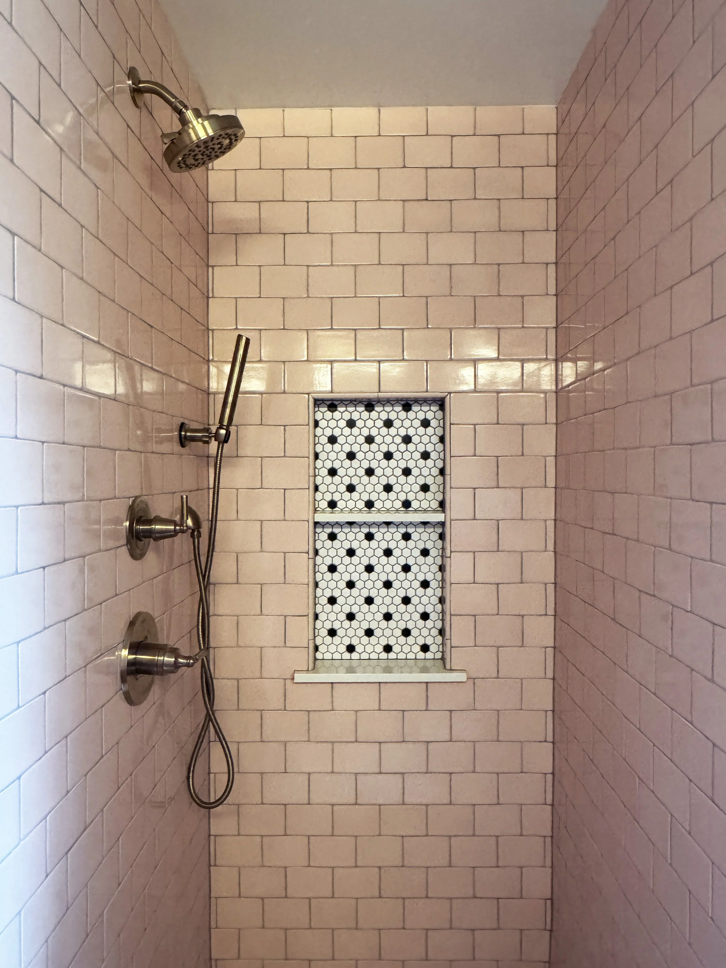 Shower with pink subway tile walls, a ceiling shower head, a handheld showerhead, and a small window with a patterned black and white curtain.
