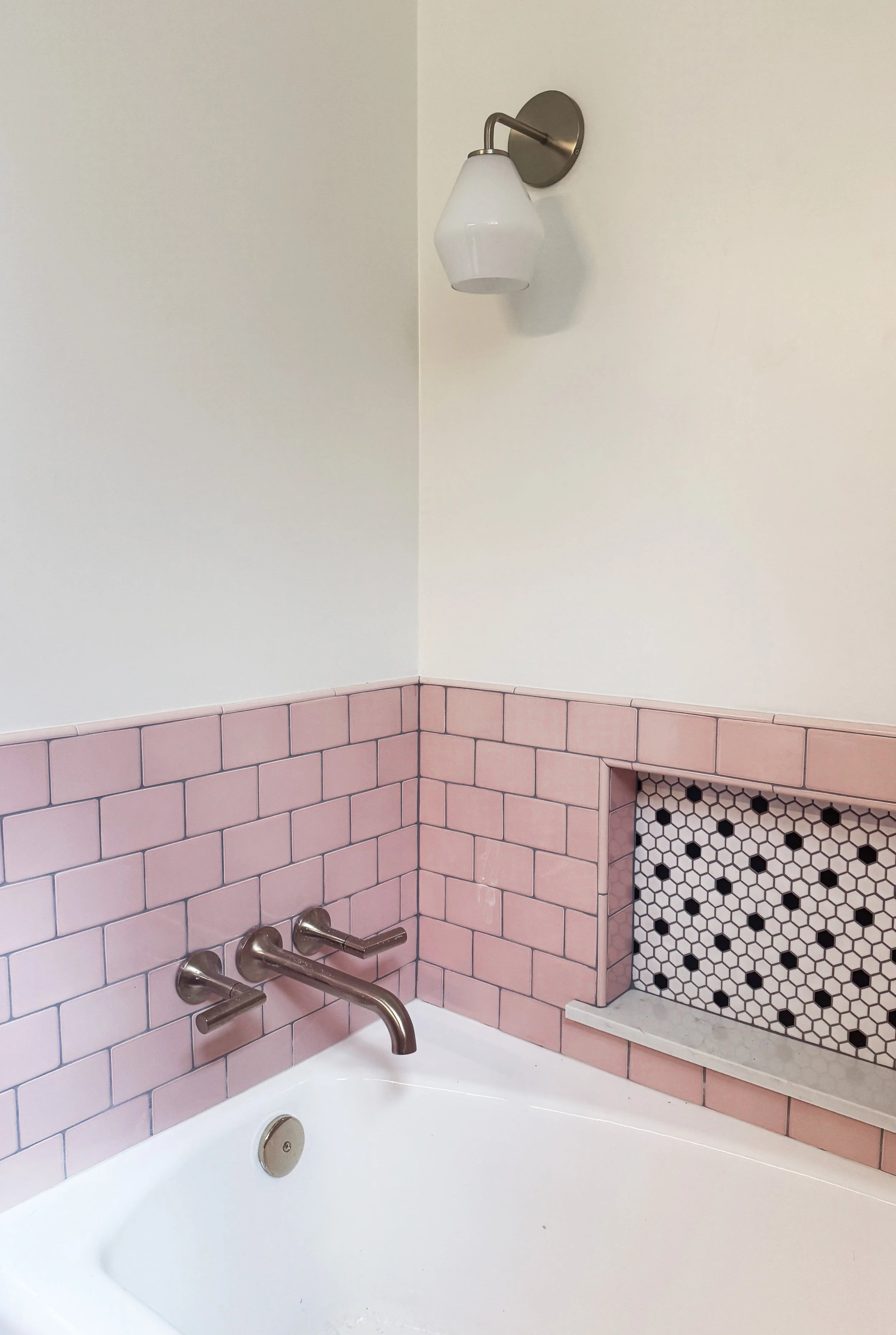 Bathroom with pink subway tile wainscoting, a built-in black and white hexagon tile shelf, white bathtub, brushed metal faucet, and a wall-mounted white glass light fixture.