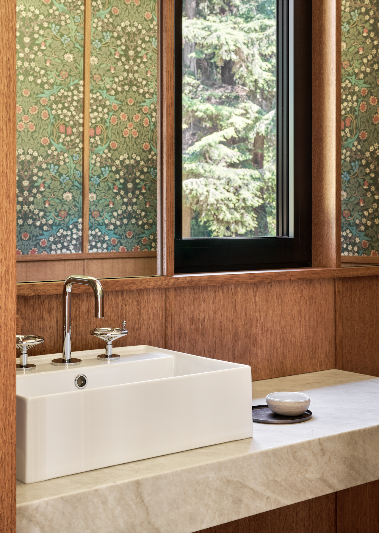 Bathroom with a white ceramic sink, a chrome faucet, a small bowl, and a window revealing green trees outside, with floral patterned wallpaper.