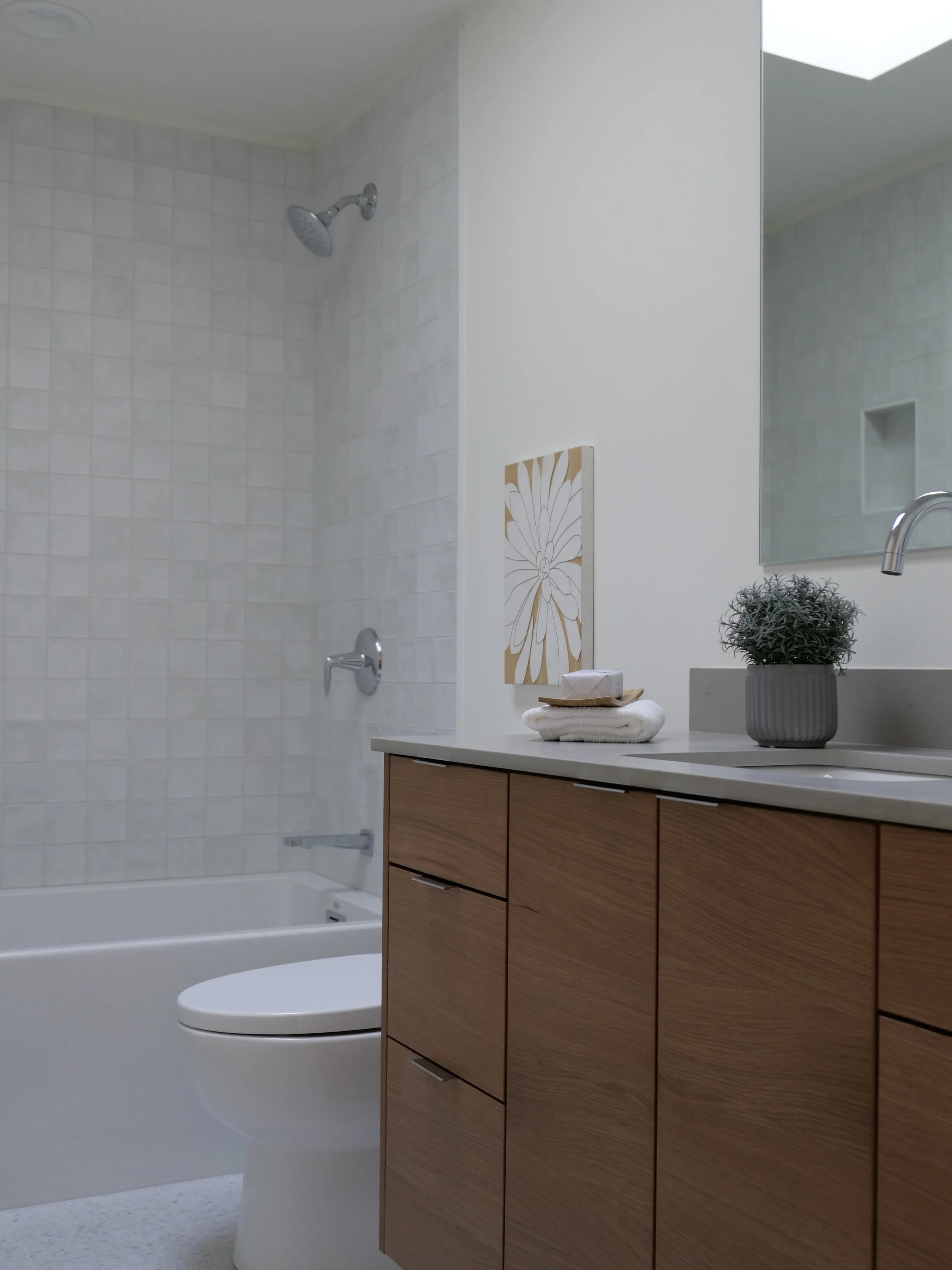 Modern bathroom with wooden vanity, potted plant, towel, and artwork, shower with tiled wall and showerhead