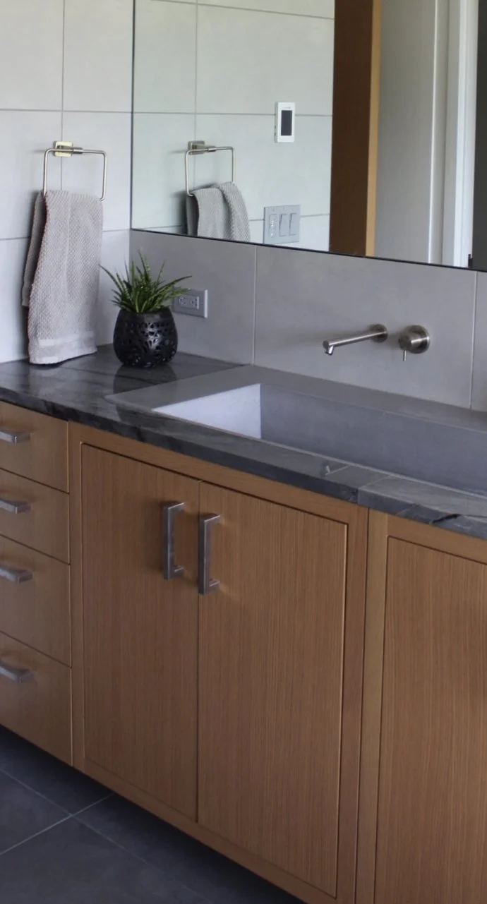 Bathroom vanity with a wooden cabinet, a gray countertop, a rectangular sink, a small black plant pot with a green plant, a towel on a wall hook, a mirror reflecting part of the room, and a wall-mounted faucet.