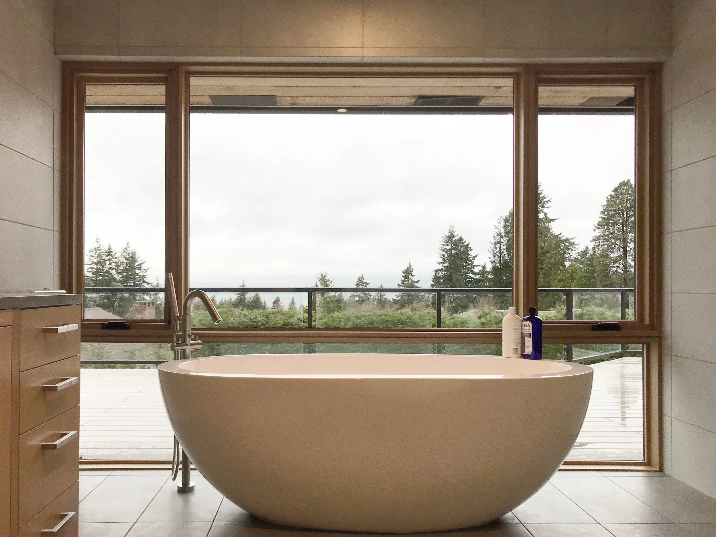Modern bathroom with a freestanding bathtub positioned in front of large glass sliding doors that open to a deck and overlook a forested landscape on a cloudy day.