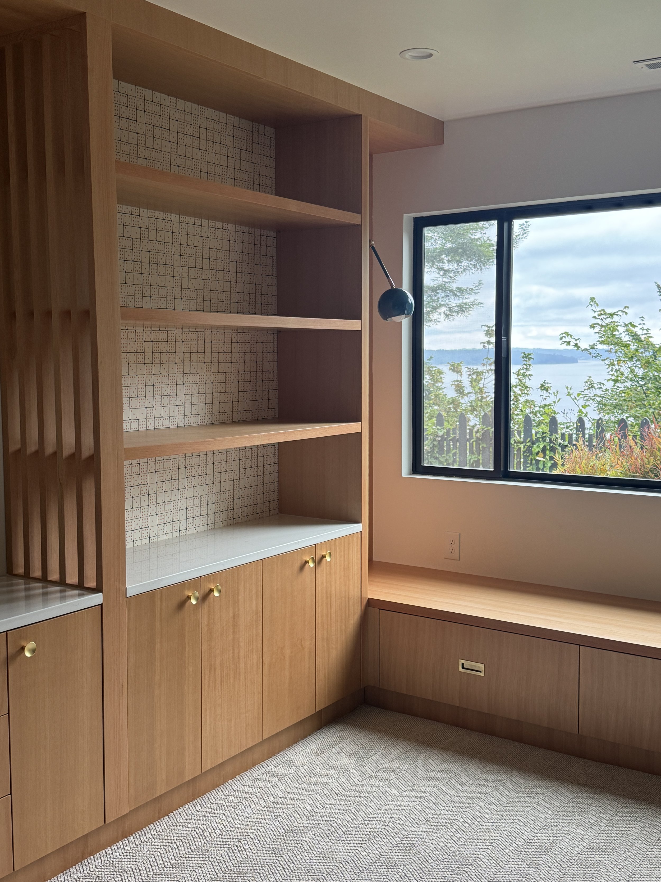 Empty wooden built-in shelves and cabinets in a room with a large window showing outdoor trees and water