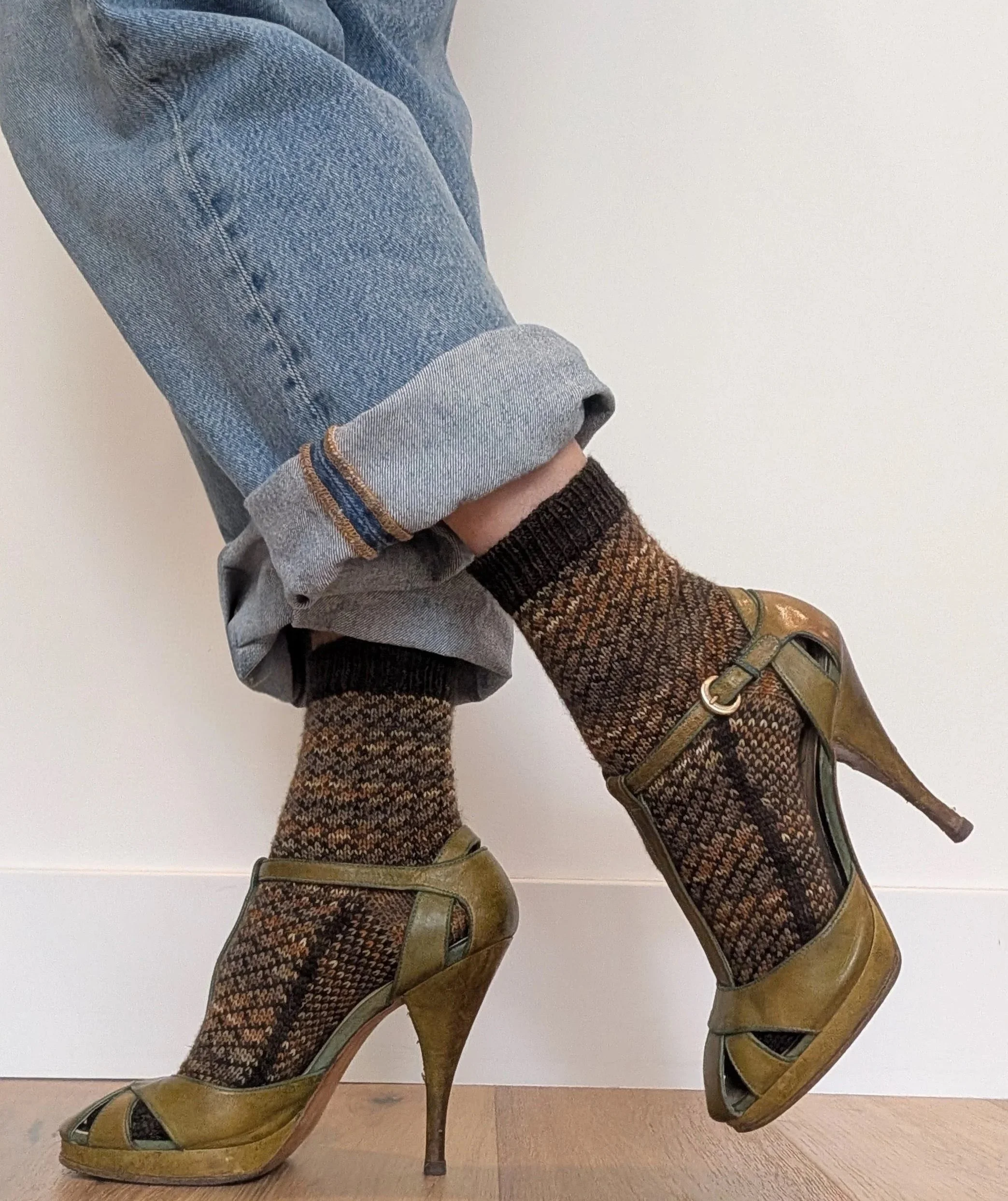 two feet are shown standing in front of a white wall, dressed in hand knit socks and green high heels.
