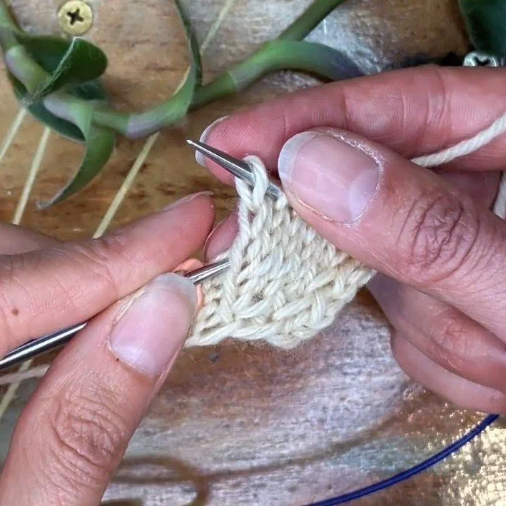 two hands hold a swatch of fabric with the left knitting needle indicating which stitch to pick up for a left lifted increase