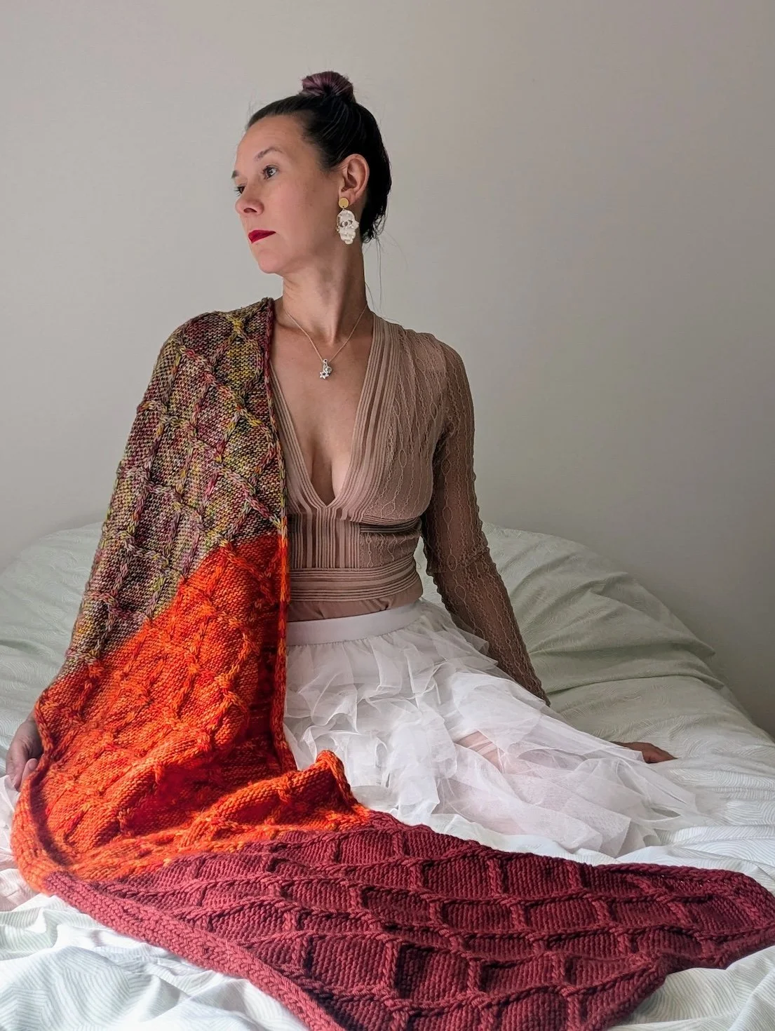 A model sits on a white bed wearing a white tulle skirt with a handknit cabled shawl running over her.